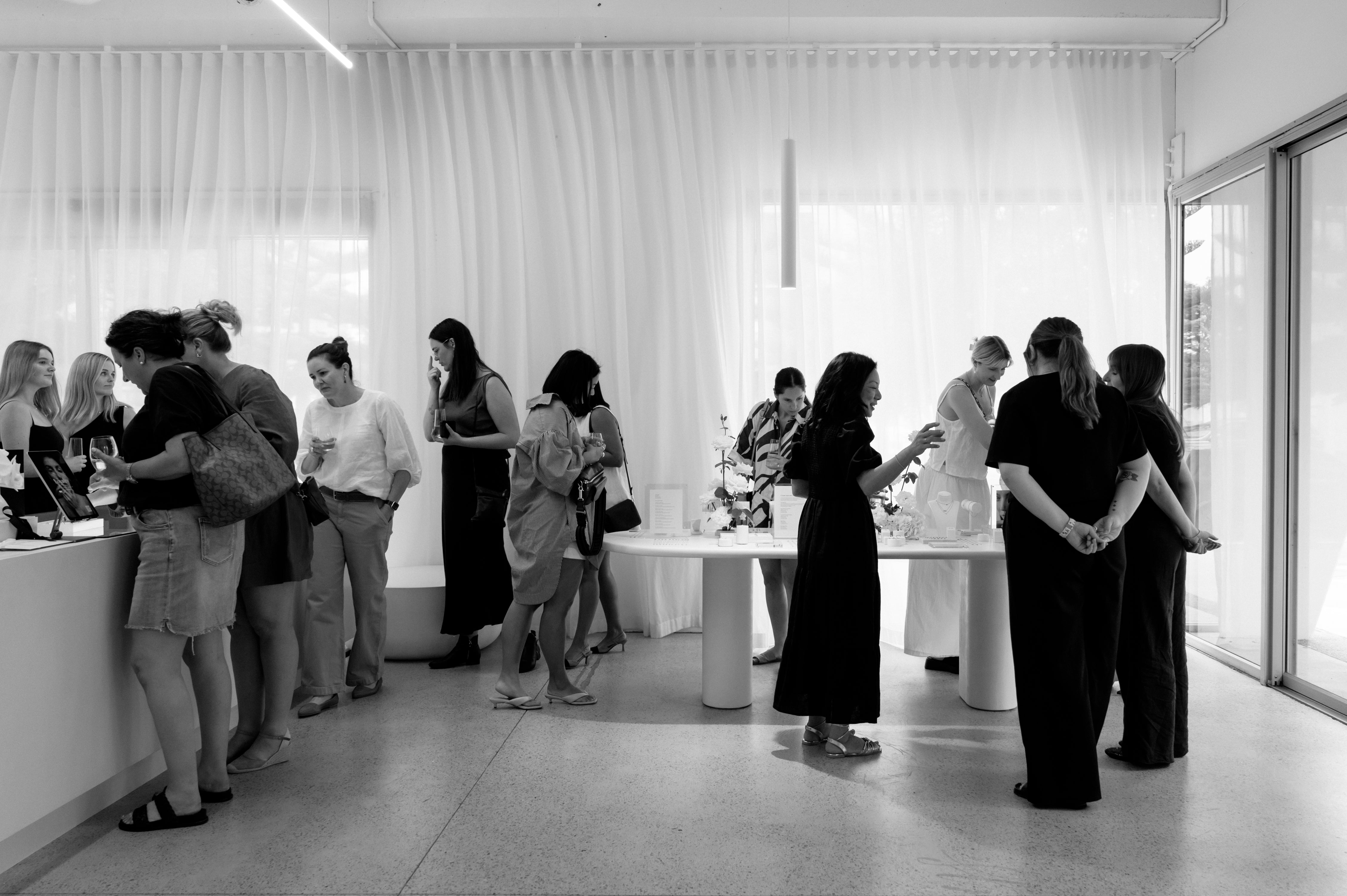 People gather in a chic, modern jewellery boutique, featuring elegant displays and soft overhead lighting against sheer curtains and sleek interiors.