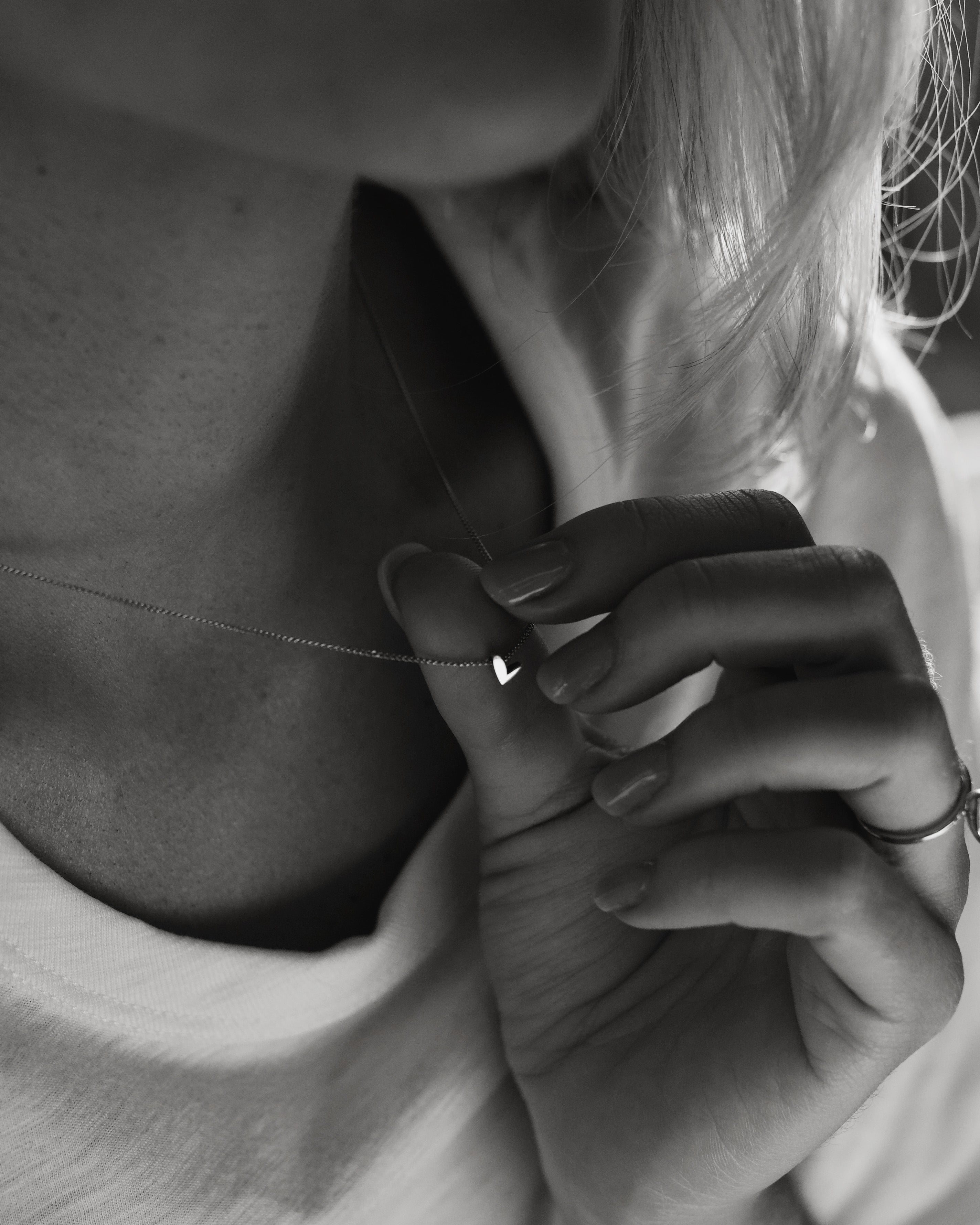 Close-up of a person wearing a delicate, high-end necklace with a small heart pendant, gently held between fingers, in soft, monochrome lighting.