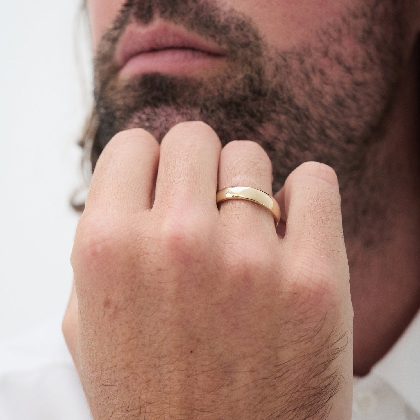 Man wearing a gold wedding band on his finger, close-up of hand near his bearded chin. Elegant, luxury jewellery showcased. [text overlay: 5mm Polished Finish in Yellow Gold]