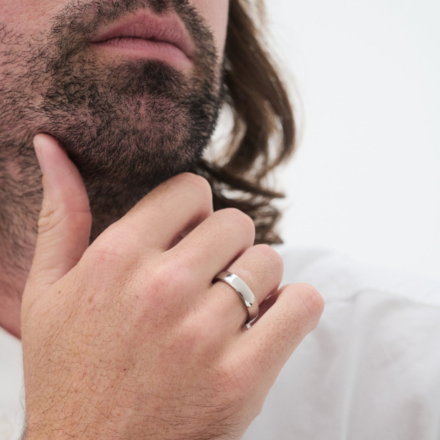 Man with a beard wearing a silver luxury ring, touching his chin thoughtfully. White background enhances the jewellery detail. [text overlay: 5mm Polished Finish in Platinum]