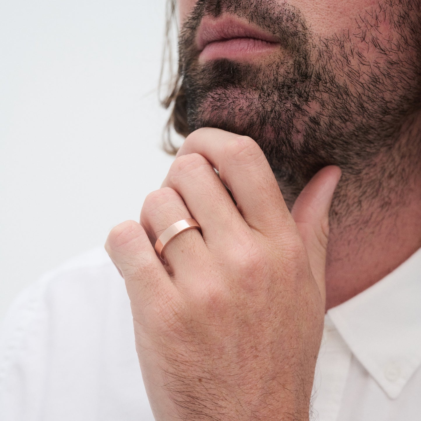 Man with beard wearing a white shirt, showcasing a sleek gold ring on his finger, touching his chin thoughtfully.