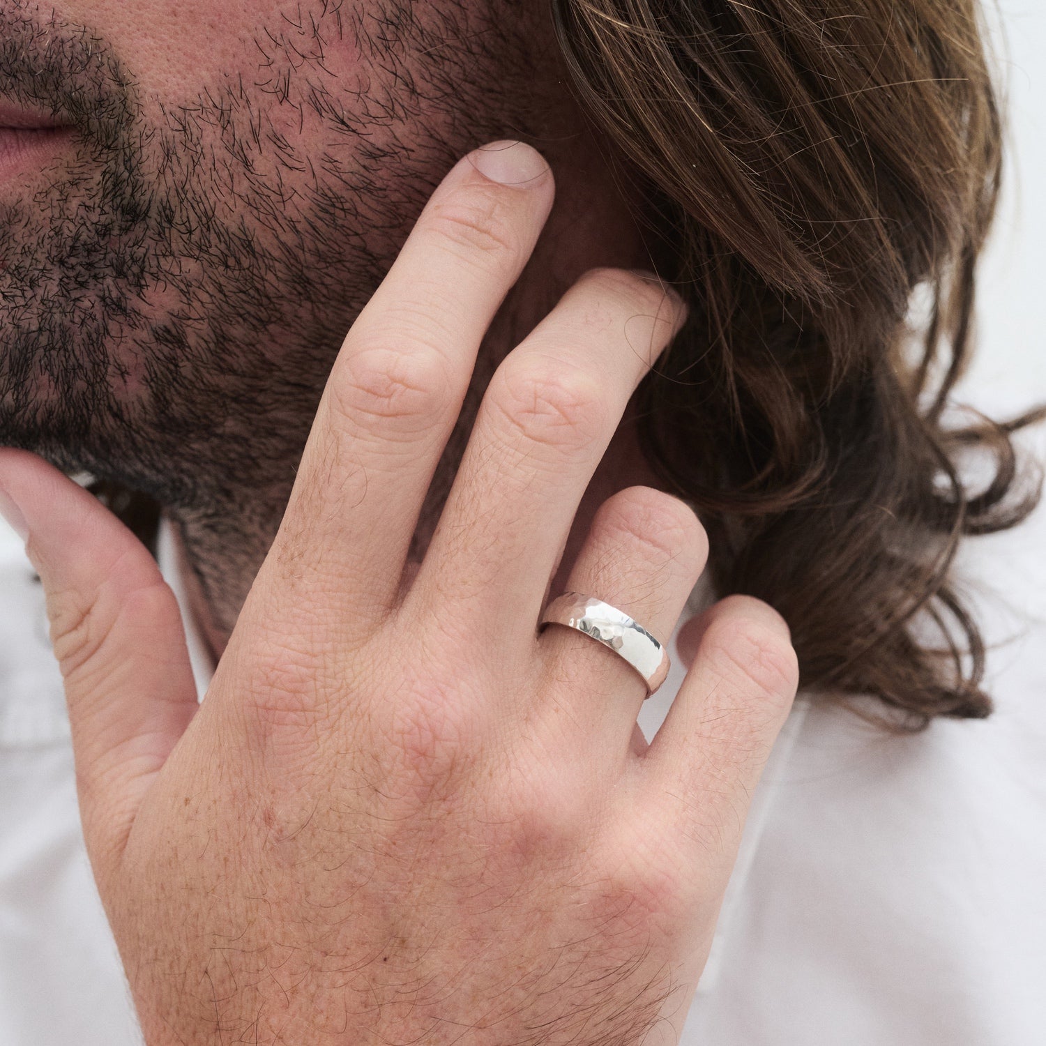Man with long hair showing a silver ring on his finger, wearing a white shirt. Focus on the ring, suggesting luxury and elegance. [text overlay: 6mm Hammered Finish in Silver]