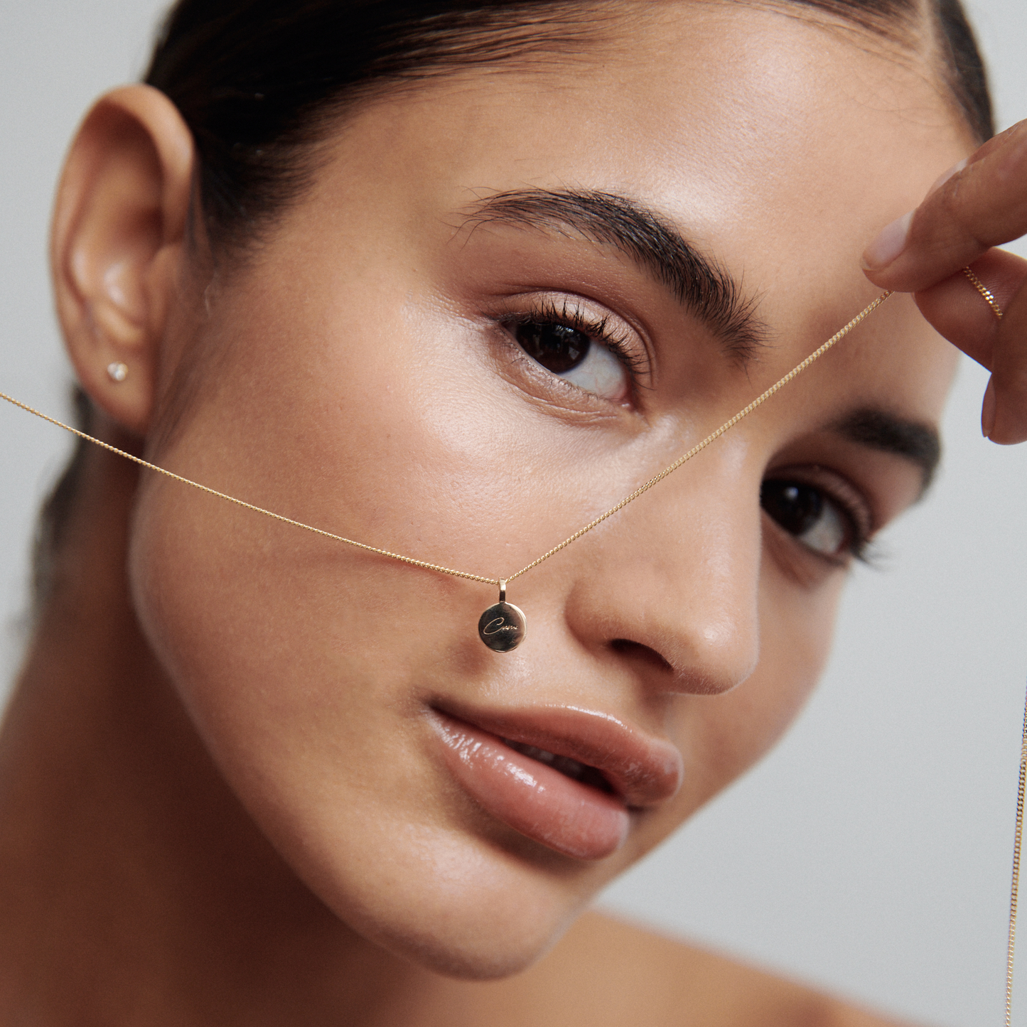 Woman holding a delicate gold necklace with a round pendant, showcasing luxury jewellery against her radiant skin, minimal makeup enhancing her features.