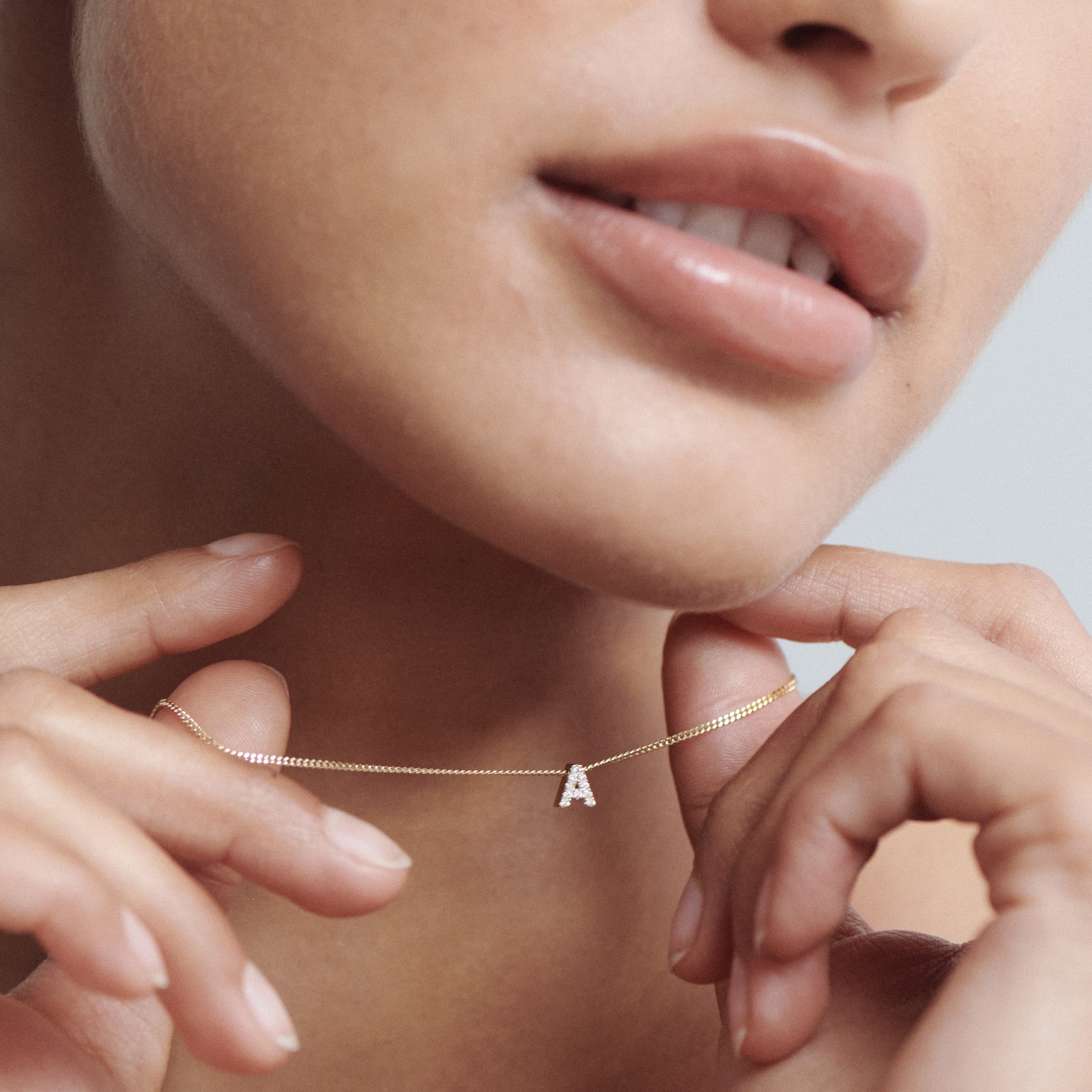 Woman holding a delicate gold necklace with a small, diamond-studded "A" pendant near her lips. Luxury jewellery close-up.