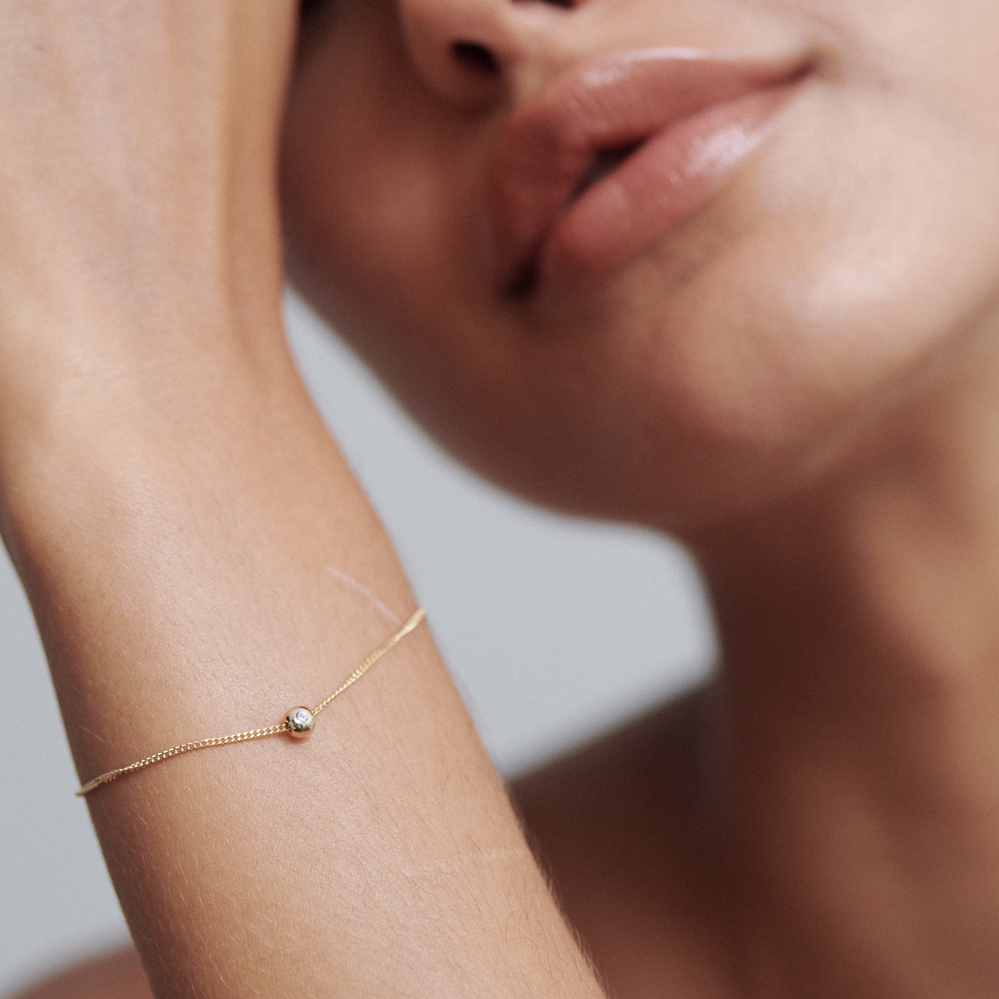 Close-up of a woman's elegant wrist adorned with a delicate gold bracelet featuring a single diamond, against a soft-focus background.