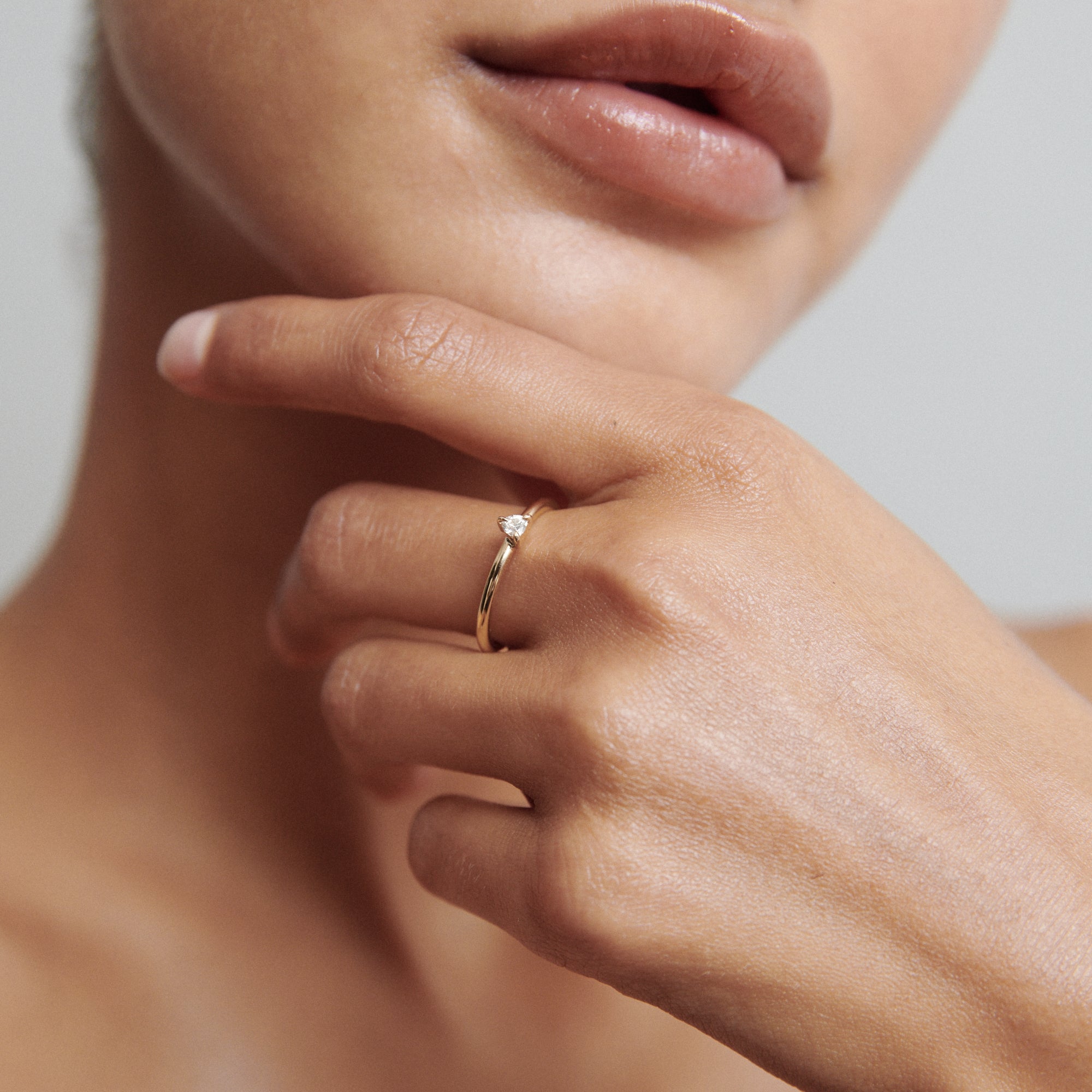 A woman's hand with a delicate gold ring featuring a small diamond, resting on her chin. Soft lighting highlights the luxury jewellery.