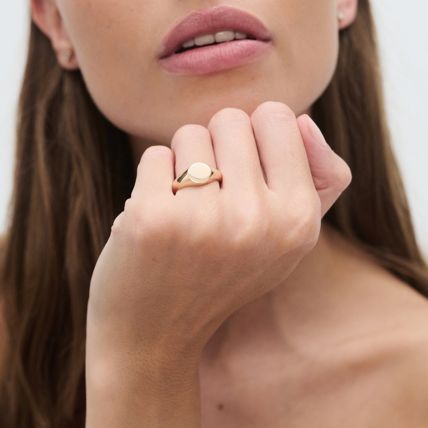A woman wearing a luxurious gold signet ring on her hand, resting near her face with light pink lips, set against a plain background.