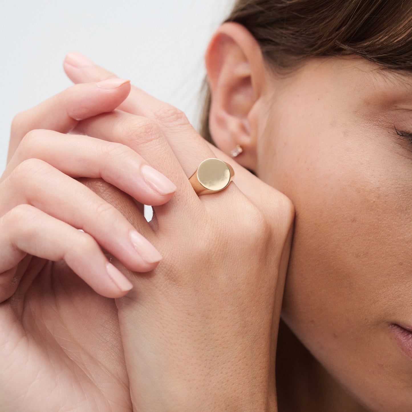 Close-up of a woman's hand with a gold signet ring on her finger, touching her face. Minimalist earrings visible. Luxury jewellery.