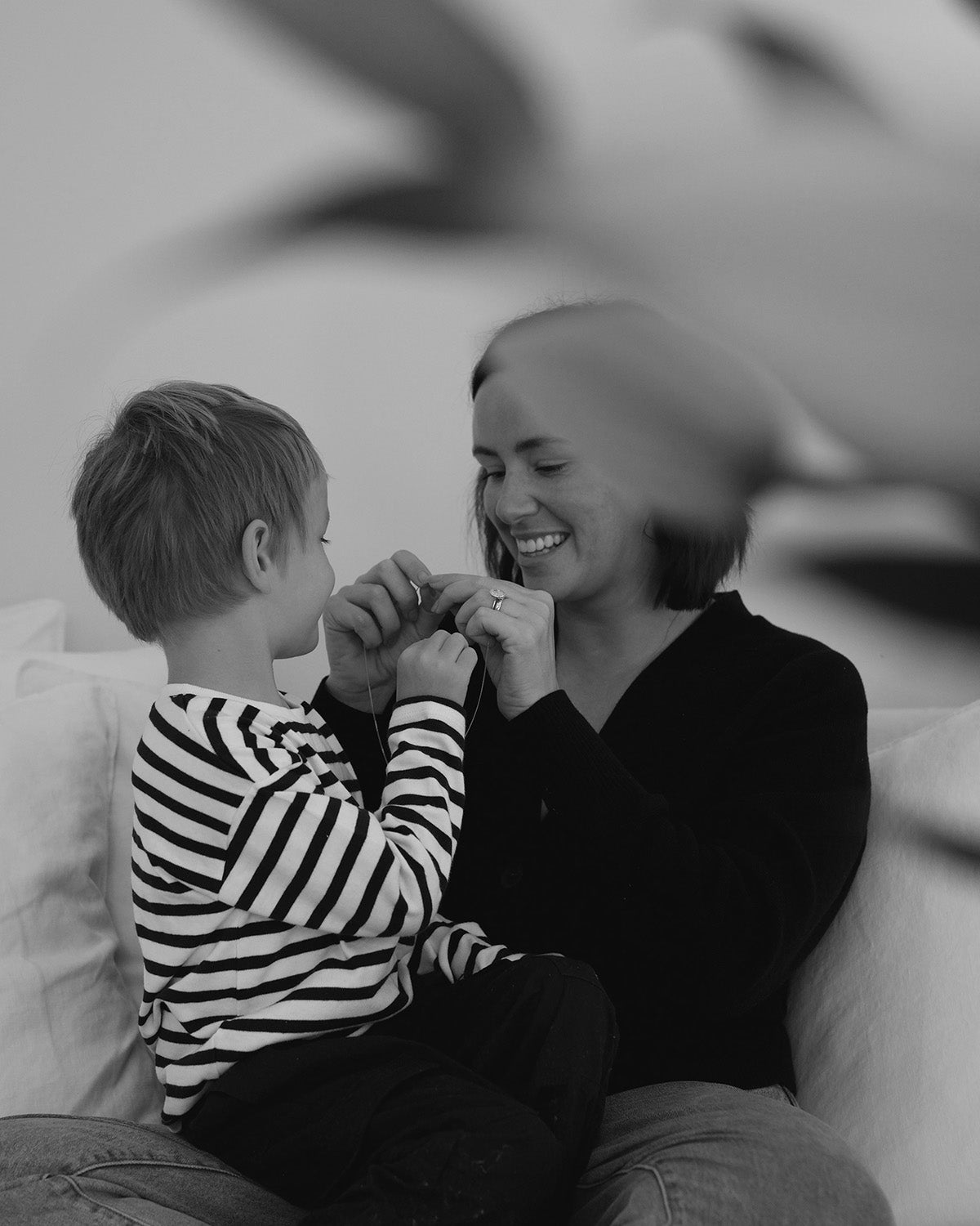 Mother and child on a couch, smiling and playing, focus on hands. Black and white image with blurred foreground leaves. Casual and warm atmosphere.