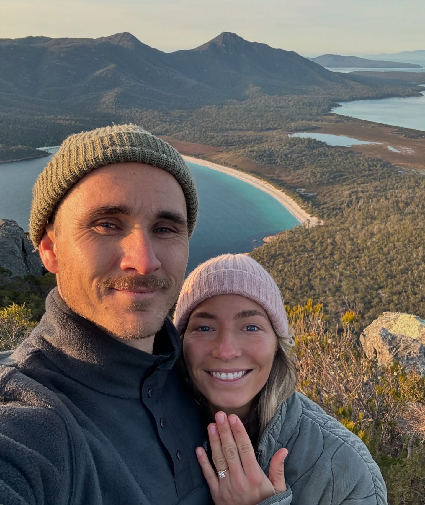 Couple stands on a mountain with coastal views; woman displays an elegant engagement ring. Scenic backdrop of ocean and mountains at sunset.