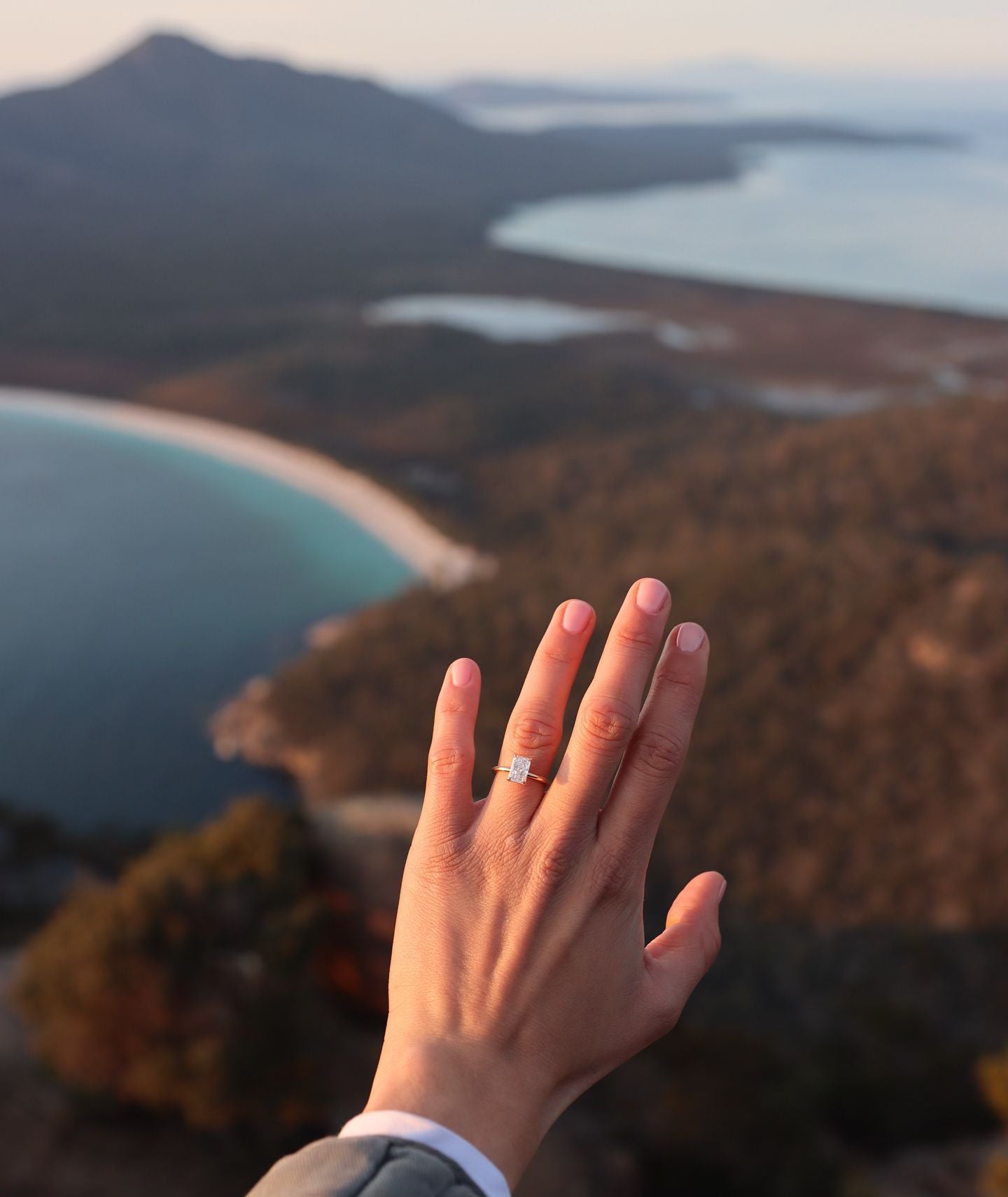 Hand displaying an elegant diamond ring with a scenic coastal backdrop of mountains and beaches. Luxury jewellery in nature setting.