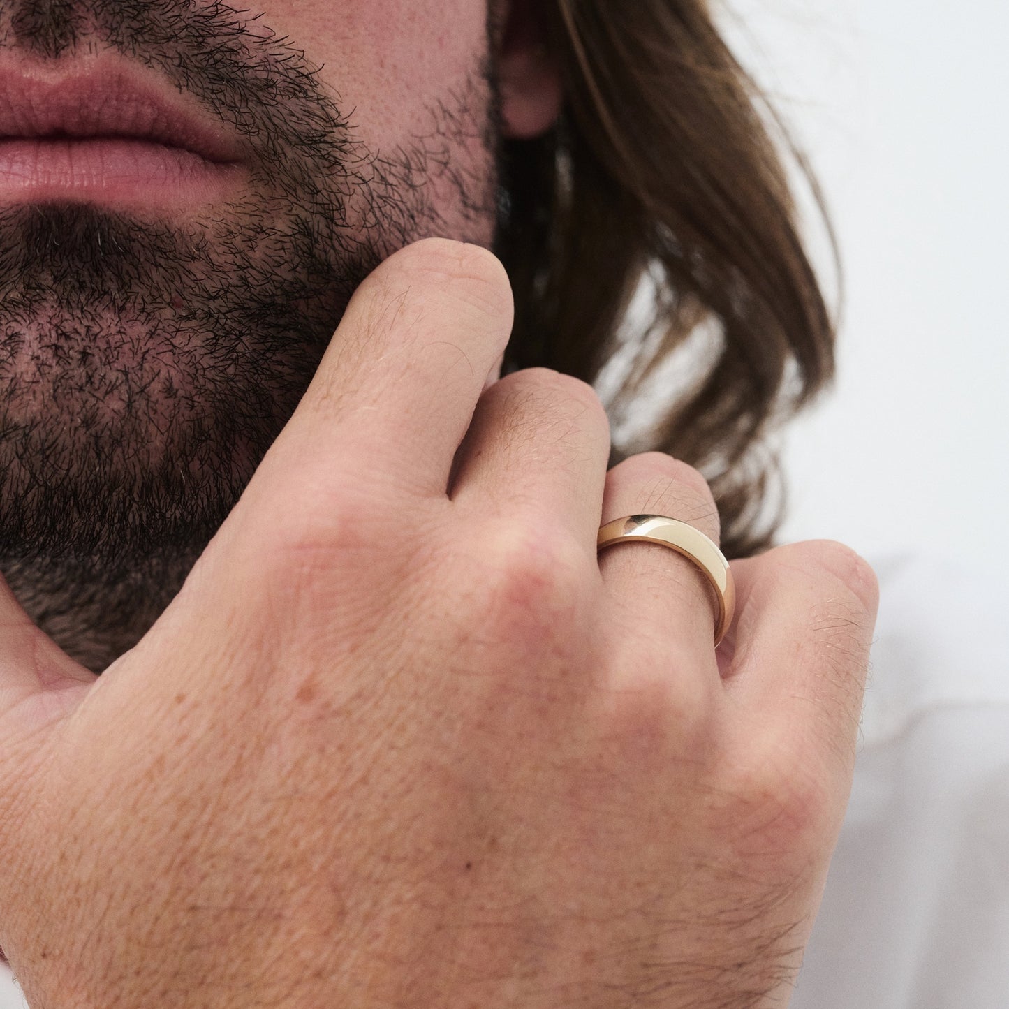 Man with a beard touching his face, wearing a polished gold ring on his finger. White background, focus on luxury jewellery. [text overlay: 6mm Polished Finish in Yellow Gold]