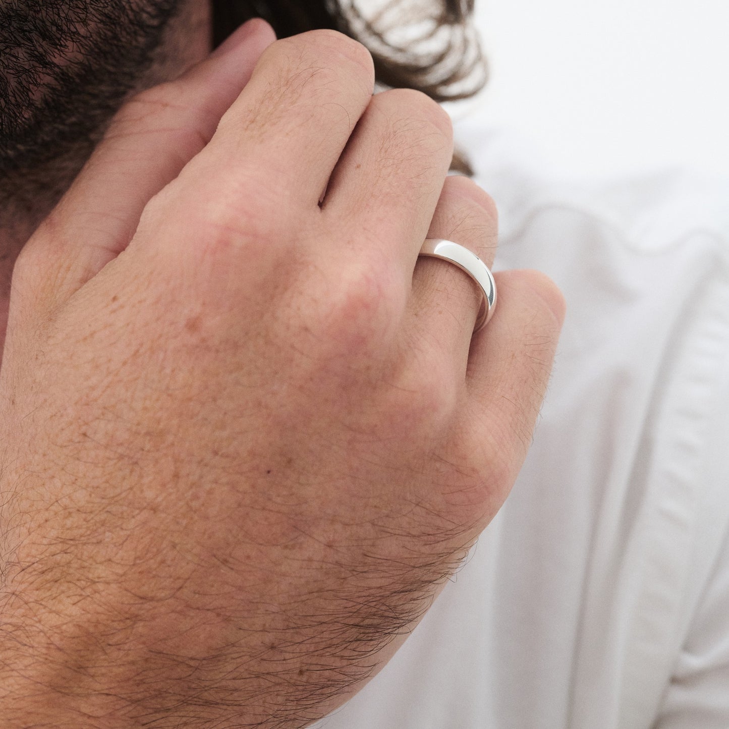 Man wearing a silver band ring, gently touching his face, dressed in white. Luxurious men's jewellery showcased with subtle elegance. [text overlay: 5mm Polished Finish in Platinum]