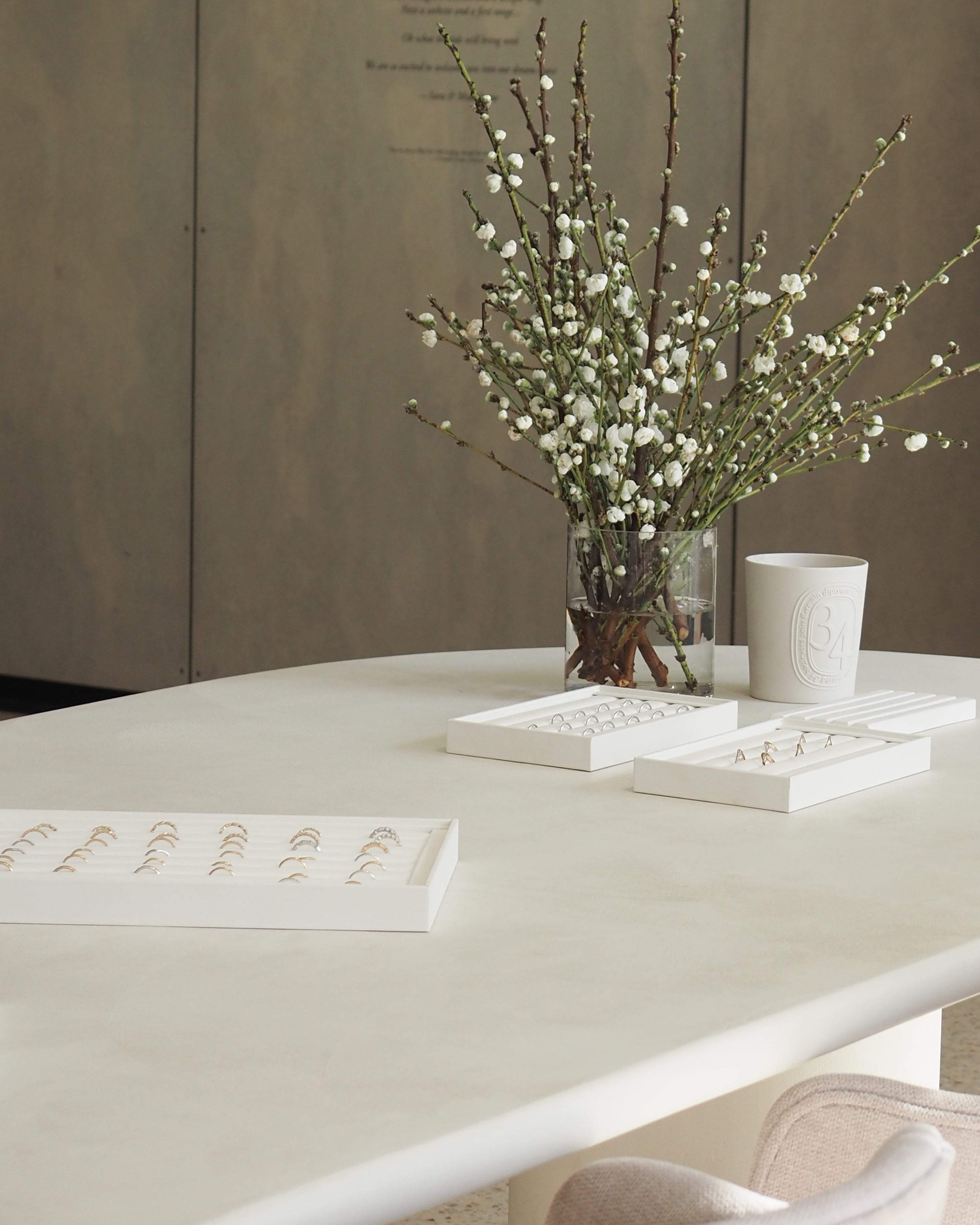 A minimalist jewellery display on a white table features gold rings and earrings in boxes; a vase with white blossoms adds elegance.