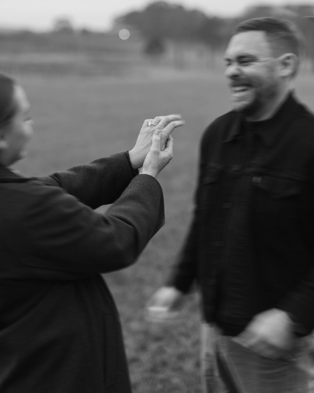 Two people in a field with woman extending a hand showing new engagement ring towards the man