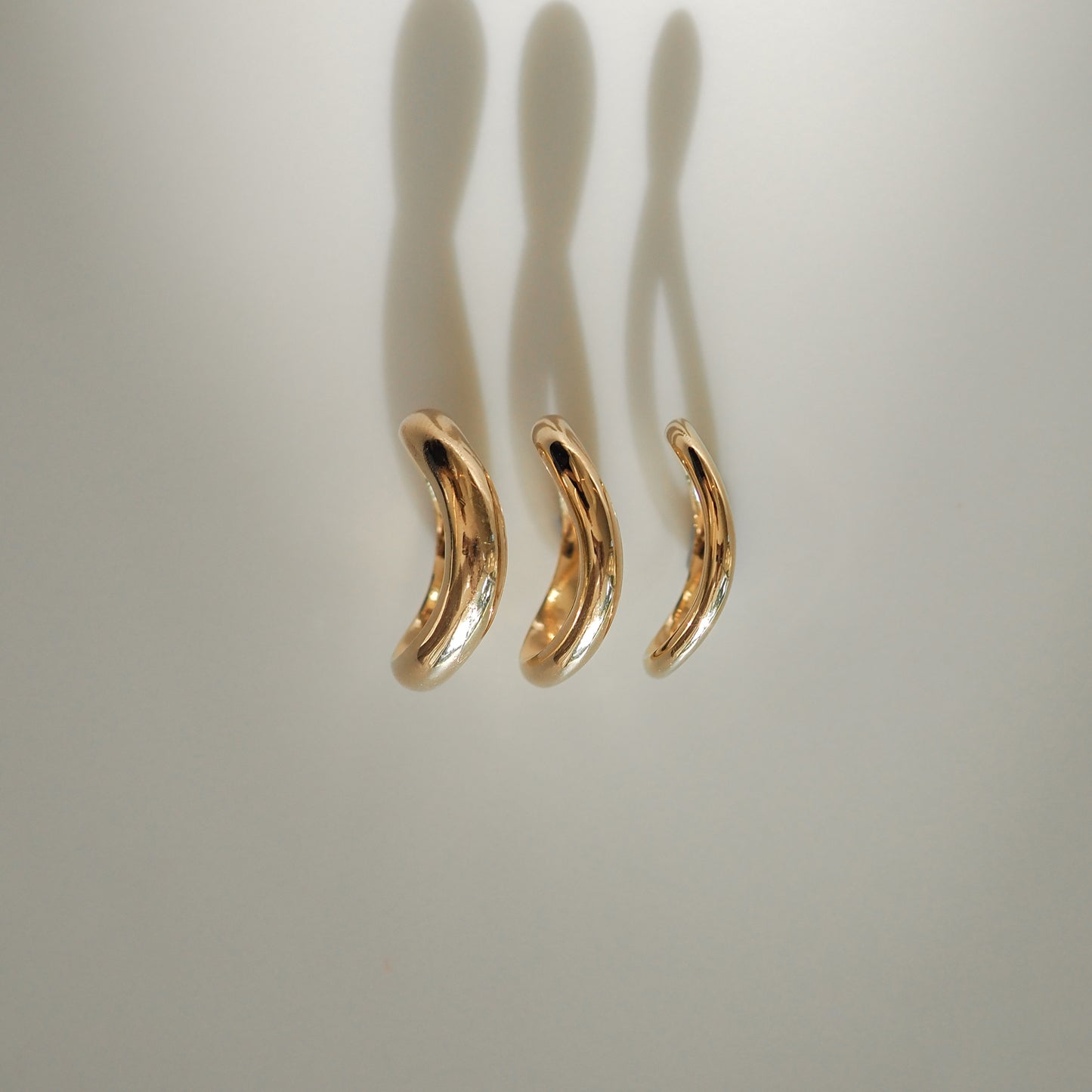 Three elegant, curved, gold earrings are arranged vertically on a light background, casting soft shadows. High-end jewellery design.