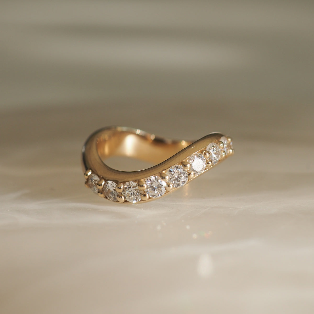 Elegant gold ring with a wave design adorned with sparkling diamonds on a soft beige background, epitomising luxury jewellery.