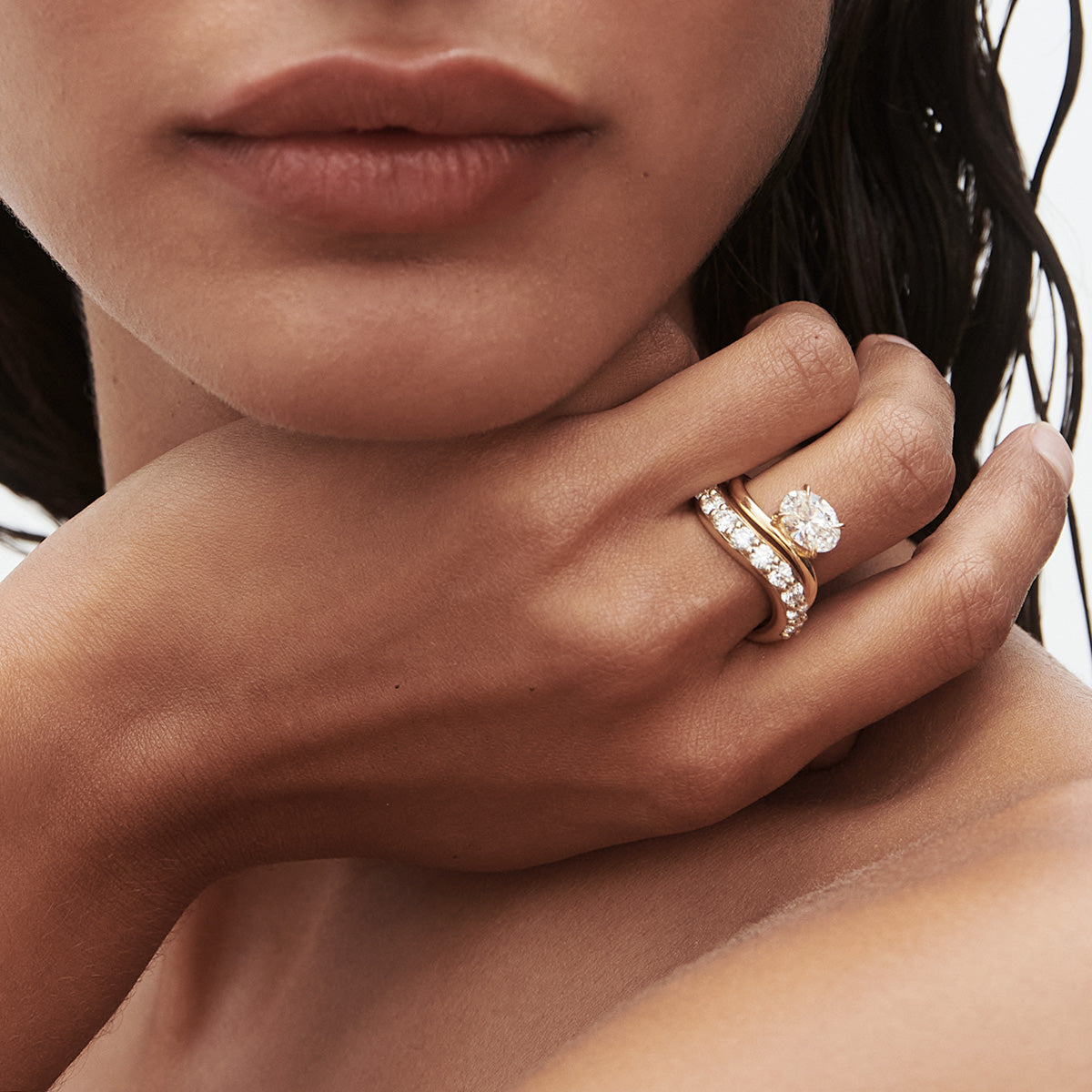 Close-up of a woman's hand adorned with a gold band and diamond ring, showcasing luxury jewellery. She gracefully touches her neck. [text overlay: Bold and Awa Oval Offset Diamond Ring] 
