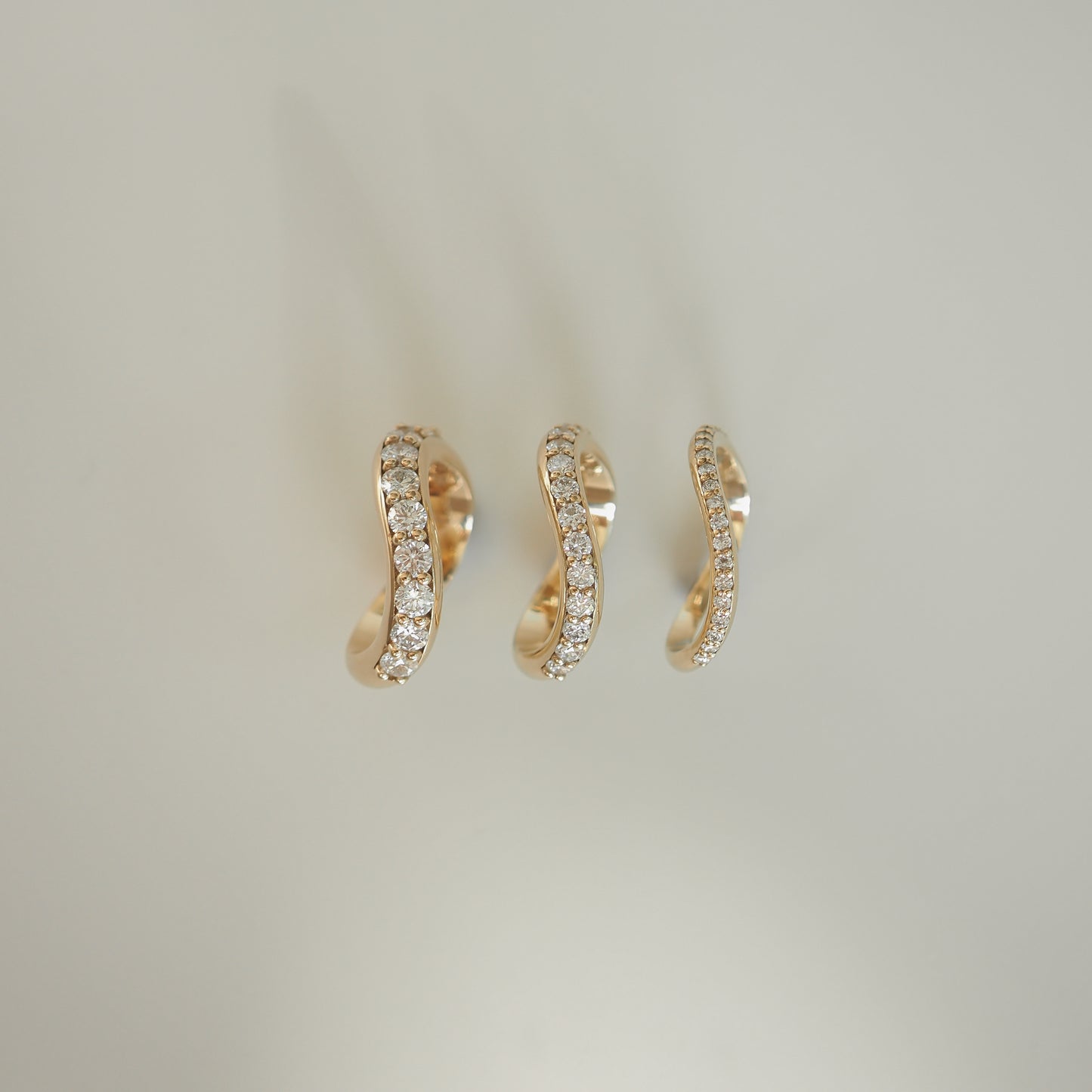 Three elegant gold hoop earrings encrusted with diamonds, arranged in a line against a soft beige background. Luxury jewellery display.