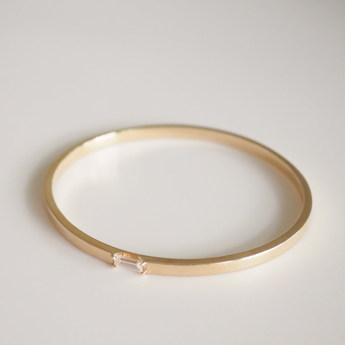 Elegant gold bangle with a small, rectangular diamond accent on a plain white background, exuding luxury and sophistication.