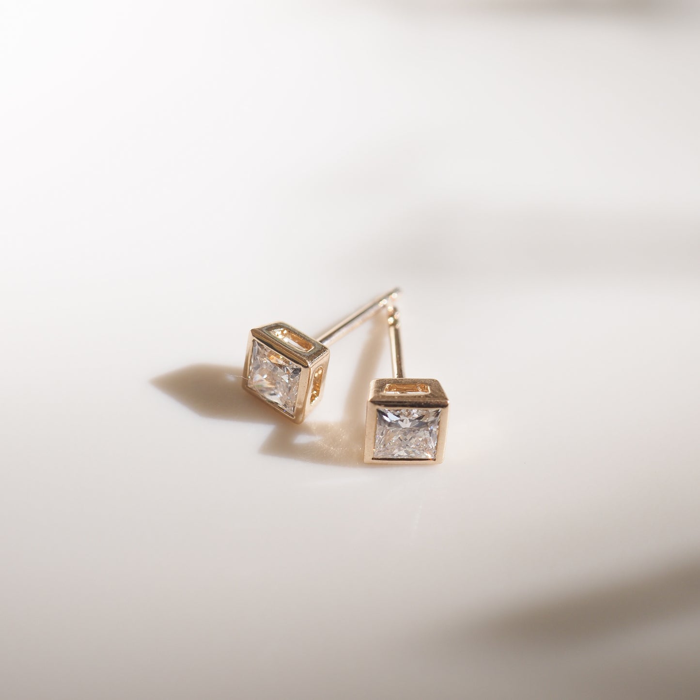 Elegant gold stud earrings with square-cut diamonds on a smooth white surface, reflecting soft light. Luxurious and minimalist jewellery.