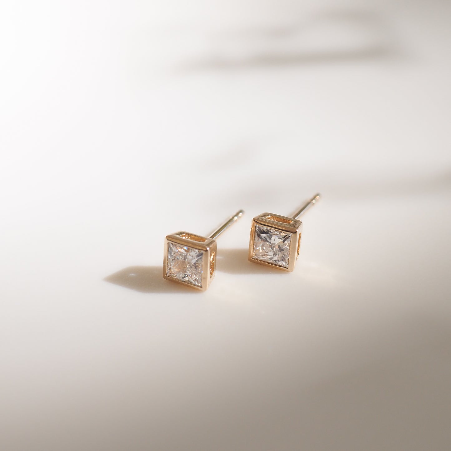 Square diamond stud earrings set in gold, elegantly displayed on a soft white surface, casting delicate shadows.