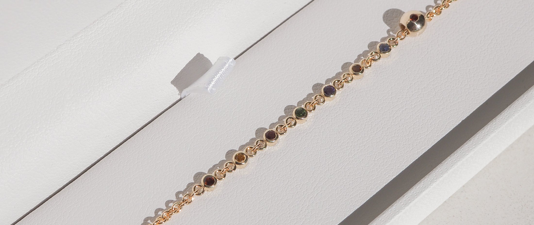 Gold bracelet with multicoloured gemstones arranged in a linear design inside a white jewellery box.