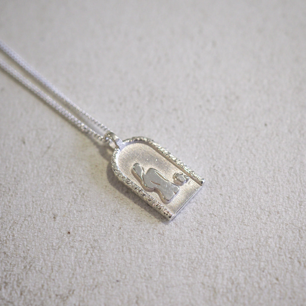 Silver arched pendant necklace on textured surface, featuring an abstract silhouette design within. Elegant and minimalist luxury jewellery.