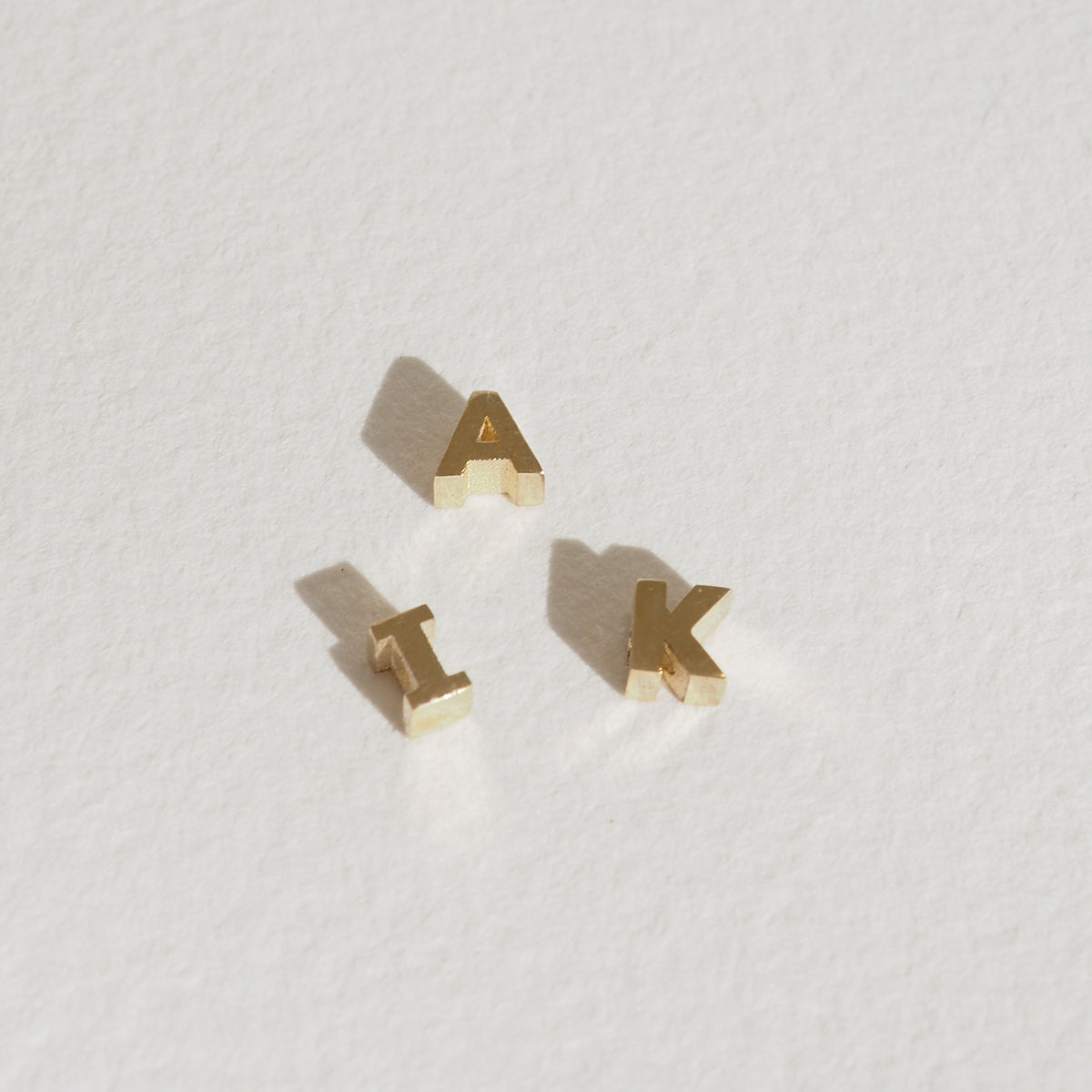 Gold letter charms "A", "I", and "K" on a textured white background, casting soft shadows. Elegant and minimalist jewellery design.