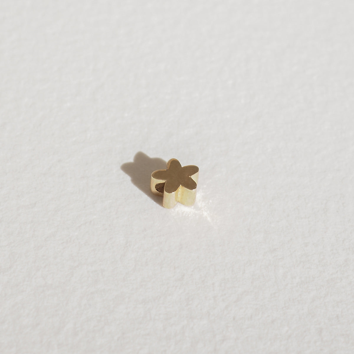 Gold clover-shaped jewellery charm on textured white surface, casting a subtle shadow. Elegant luxury jewellery piece, minimalist design.