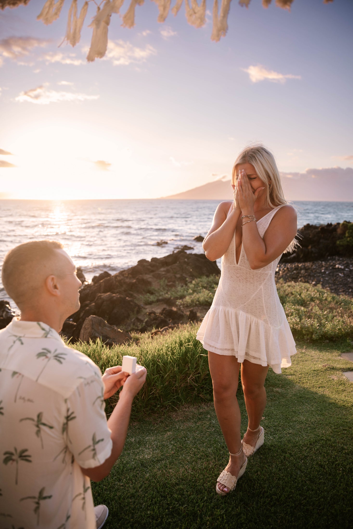 Man proposing to a woman at sunset by the ocean. She wears a white dress, covering her face with surprise. A romantic, luxury jewellery moment.