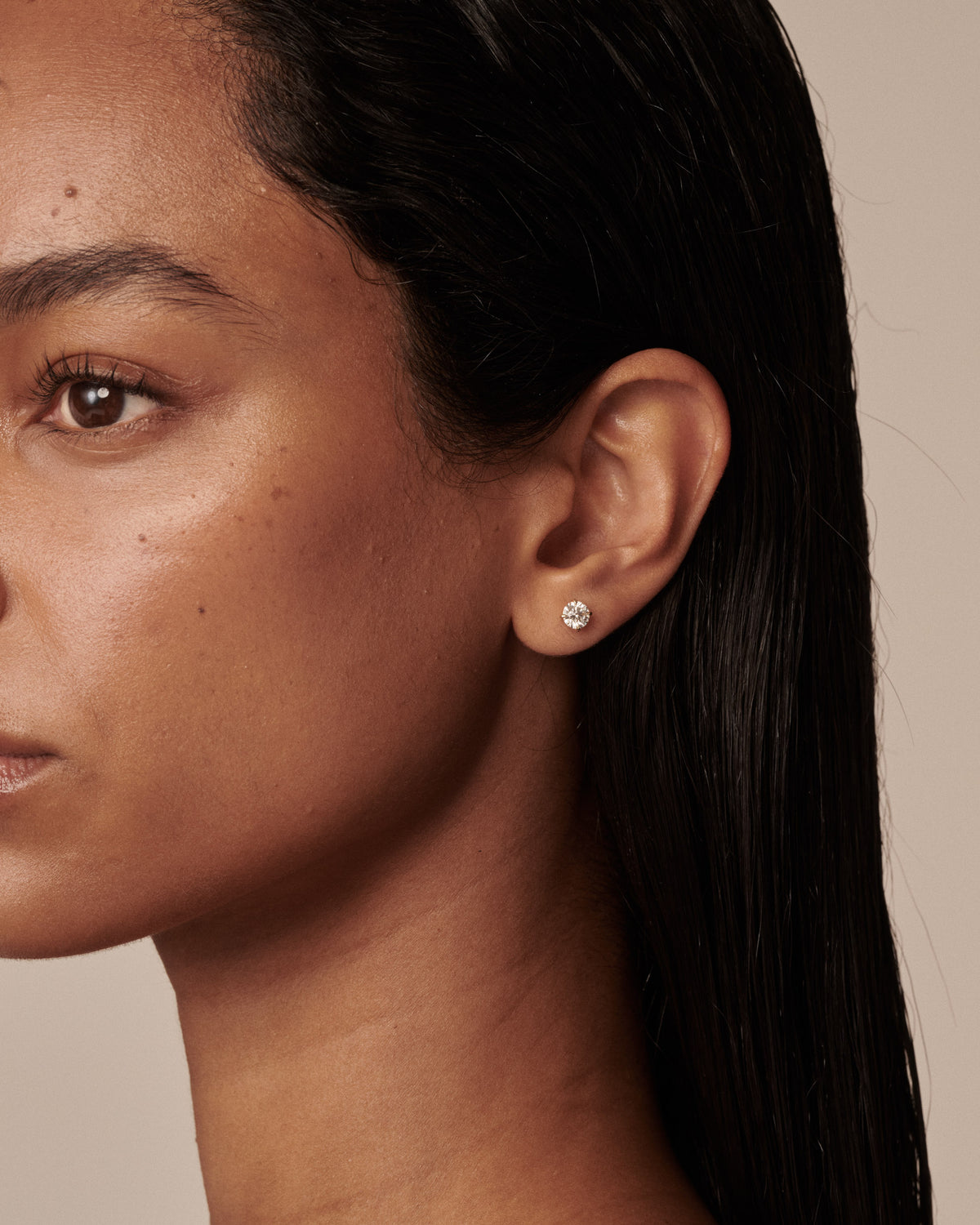 A close-up side profile of a woman with dark hair wearing a small round stud earring with a clear diamond on a warm beige background.