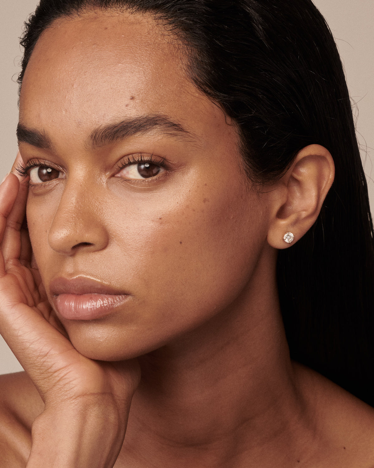 A close-up portrait of a woman with dark hair, wearing a small round silver stud earring with a clear diamond, set against a warm beige background.