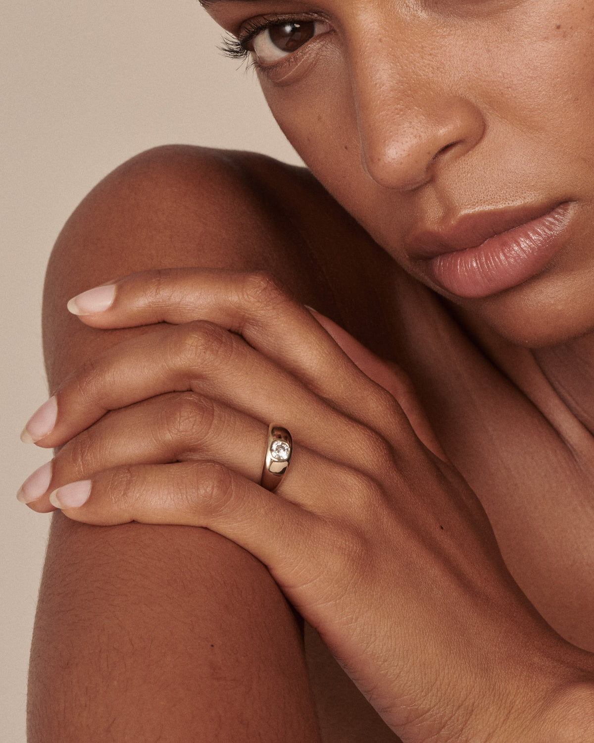 A close-up lifestyle shot of a model wearing a gold ring with a small clear diamond on her finger, resting her hand on her shoulder against a warm beige background.