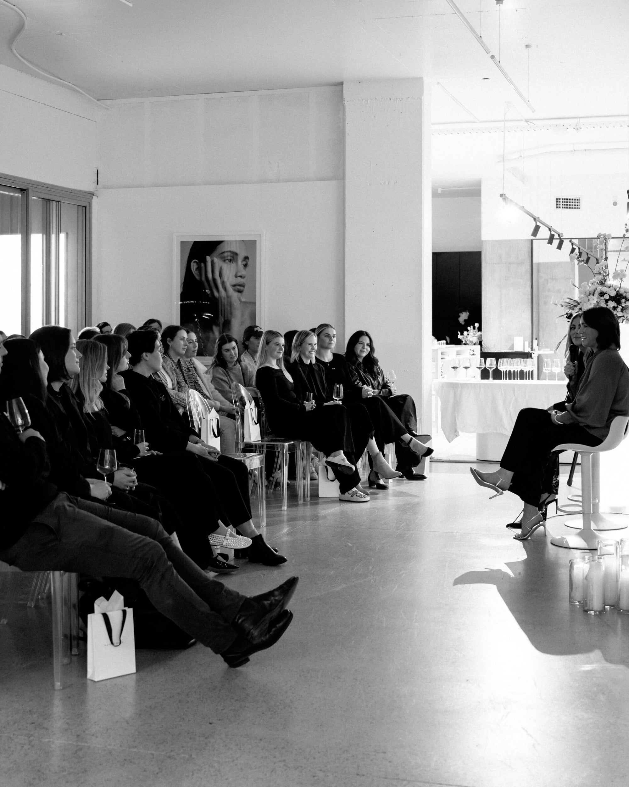 A group of people attentively listening to a speaker in a stylish, modern room with a luxury jewellery display and elegant decor.