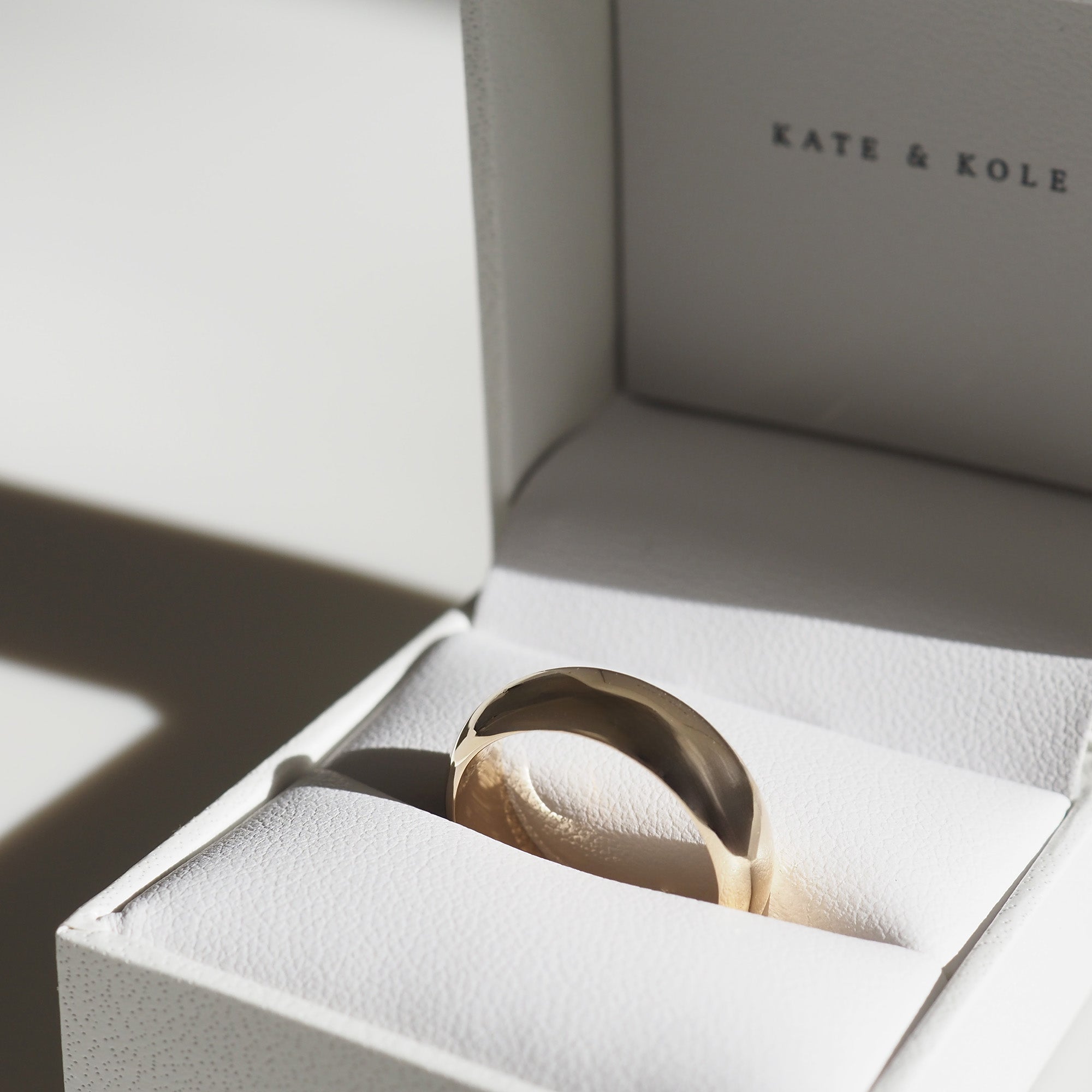 Gold ring in a white jewellery box with "Kate & Kole" text. Sunlight casts a soft shadow on the ring and box, creating a luxurious feel.
