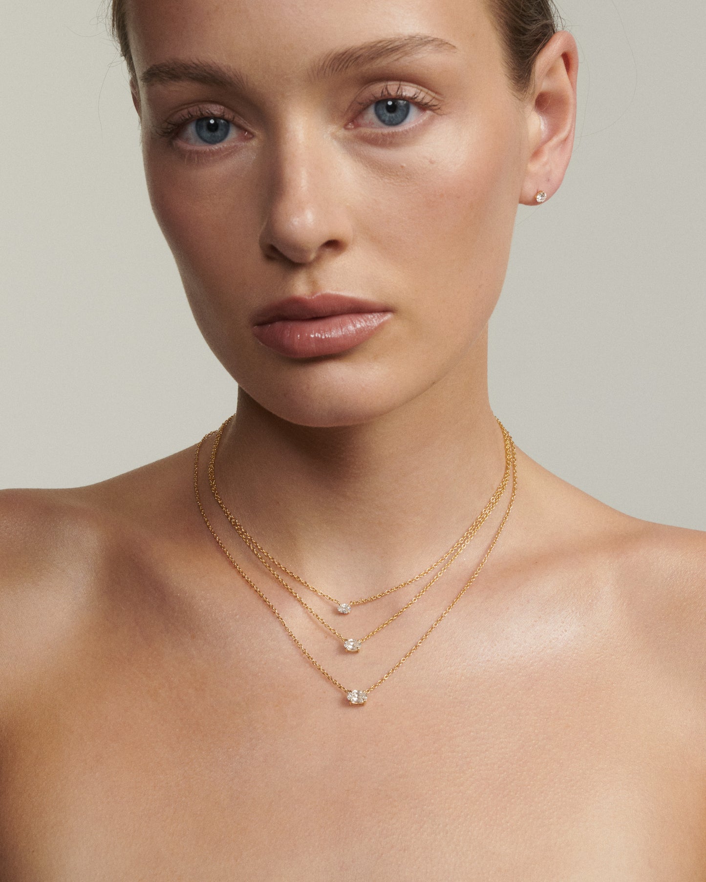 Woman wearing a delicate gold necklace with small pendants against a neutral background [text overlay: Andrea wears the 0.25, 0.5ct & 0.75ct East West Fixed Oval Diamond Necklace]