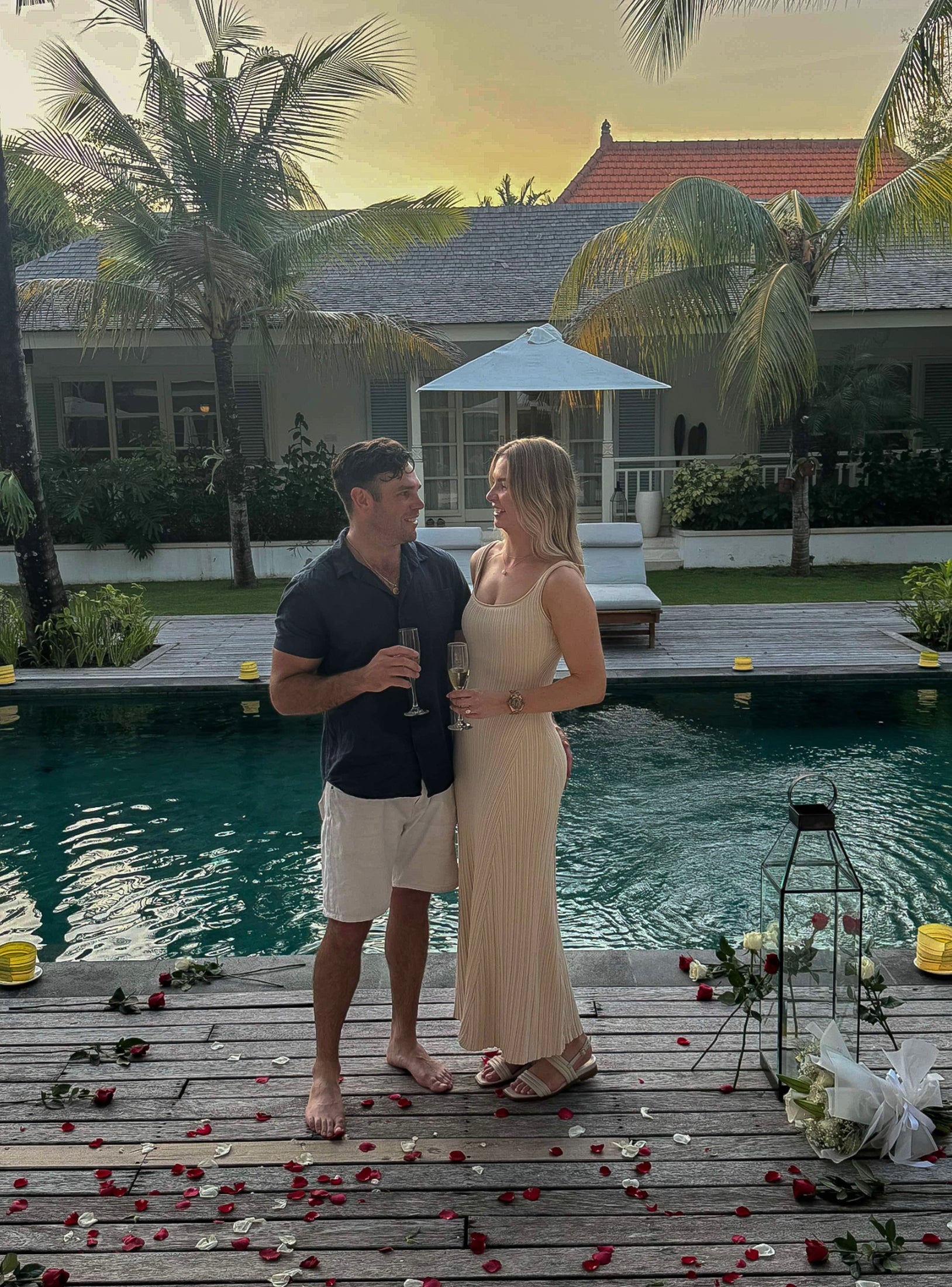 A couple by a pool at dusk, surrounded by rose petals and candles. They toast with champagne, creating a romantic luxury jewellery setting.