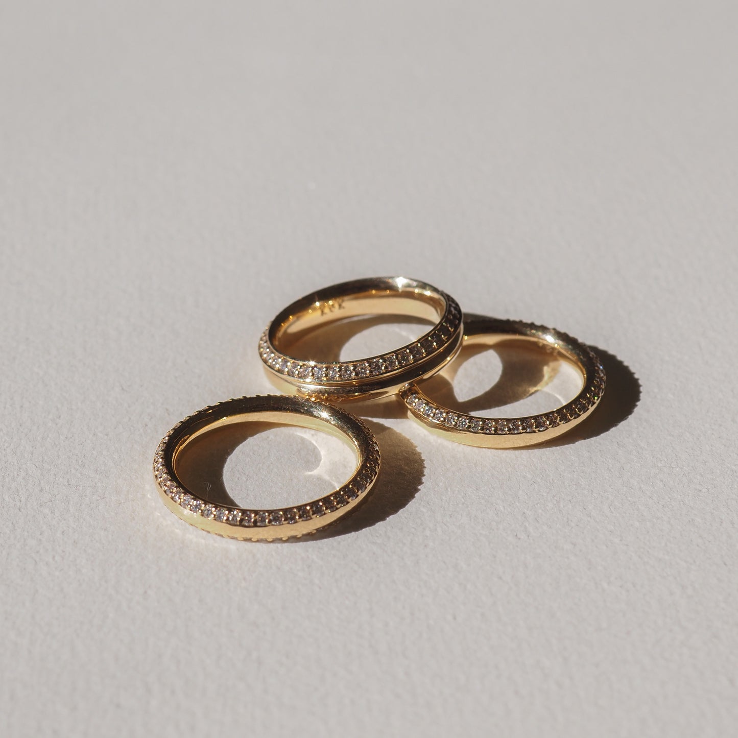 Three gold rings with small embedded diamonds arranged on a light surface, casting subtle shadows. Perfect for luxury jewellery enthusiasts.