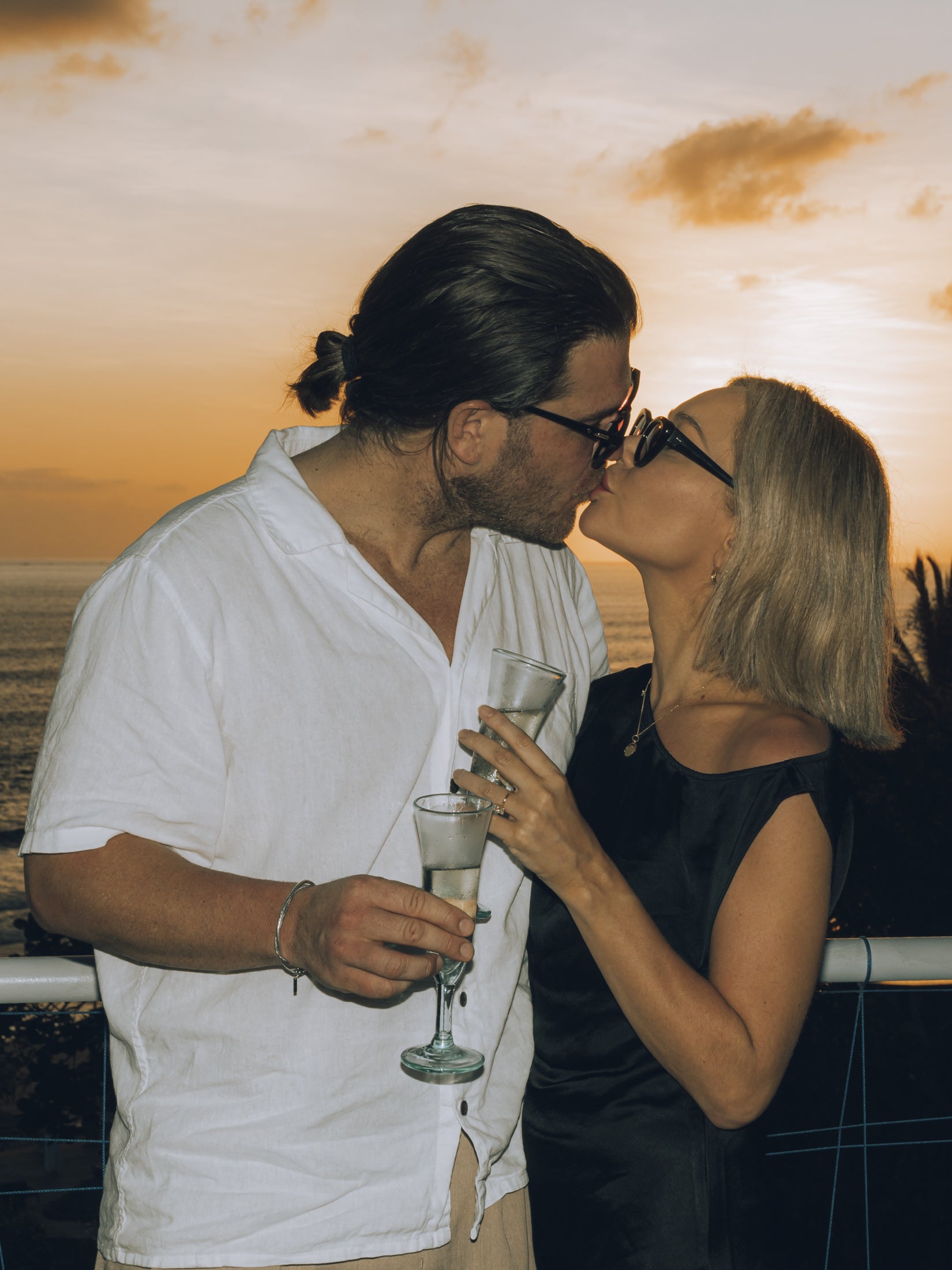 Couple shares a toast and kiss at sunset, wearing elegant attire; she sports a stylish black dress and chic sunglasses, holding champagne glasses.