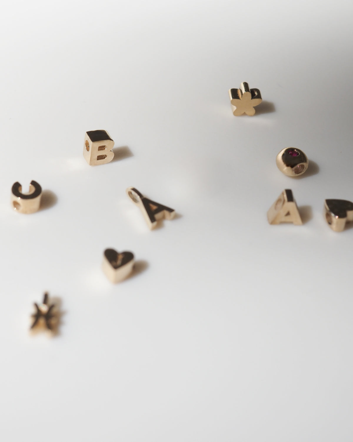 Gold letter charms scattered on a white surface, with some symbols and a small red gemstone accent.