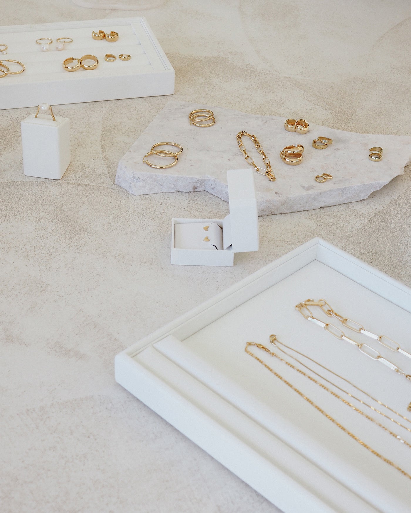 Assorted luxury gold jewellery, including necklaces, rings, and earrings, elegantly displayed on marble and white trays on a beige surface.