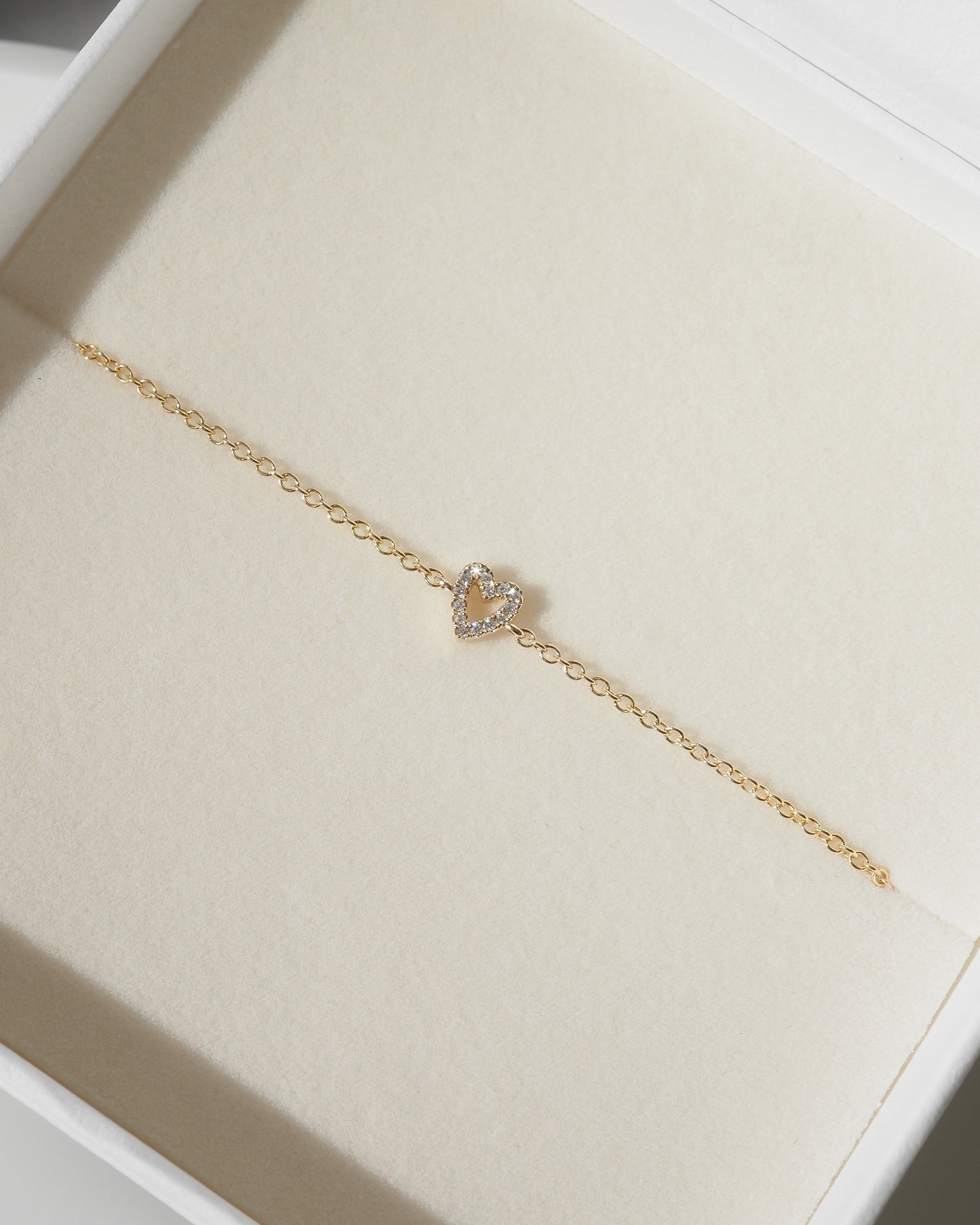 A delicate gold chain bracelet with a small heart-shaped crystal pendant, displayed inside a white jewelry box with a cream-colored lining.