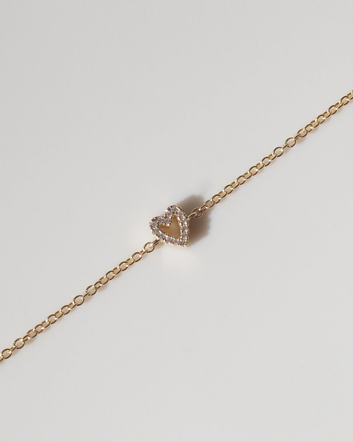 A delicate rose gold chain bracelet featuring a small heart-shaped charm with a double halo of clear stones, displayed diagonally on a light gray background.