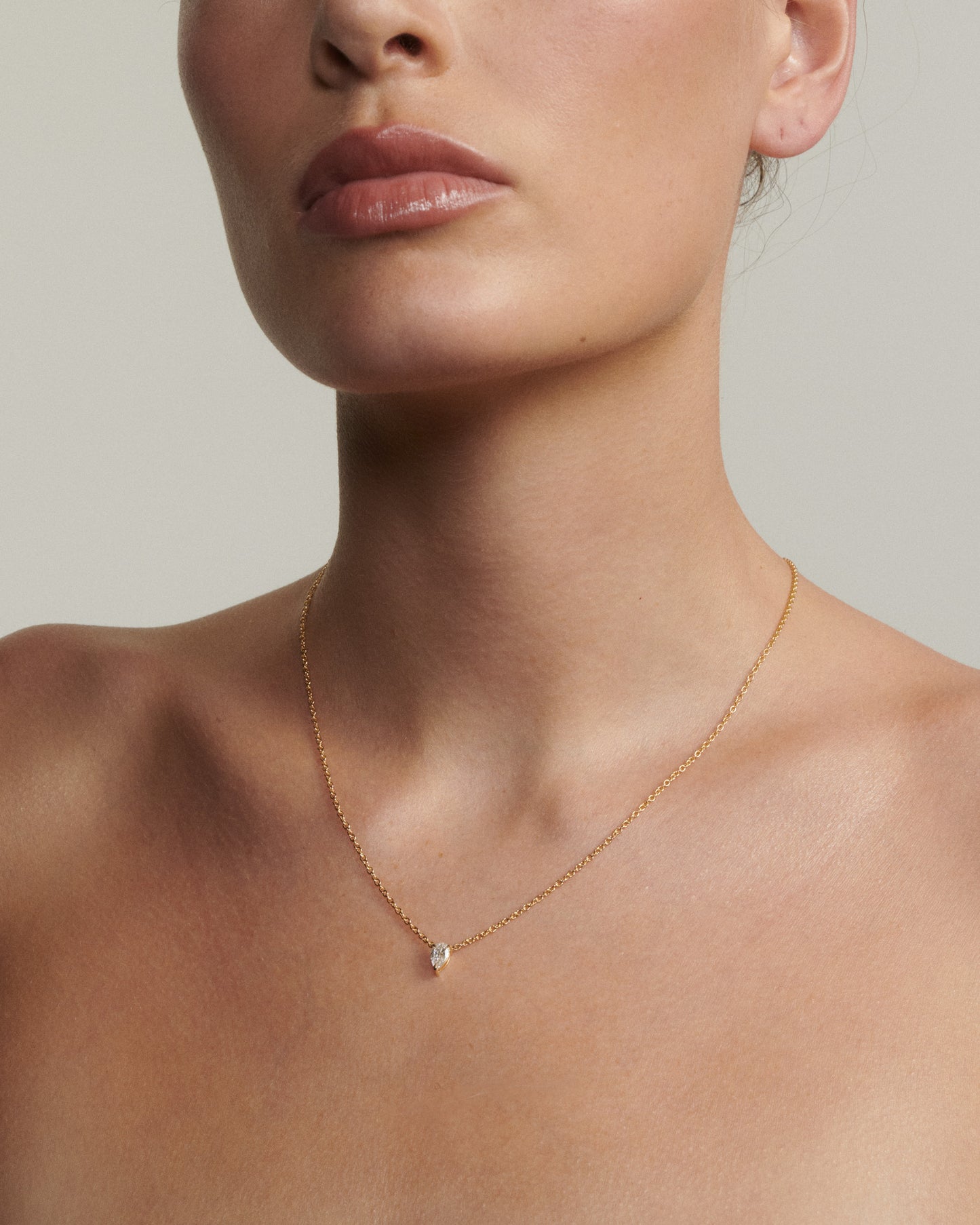 Close-up of a person wearing a delicate gold necklace with a small pendant on a neutral background [text overlay: Andrea wears the 0.5ct Pear Diamond Necklace]