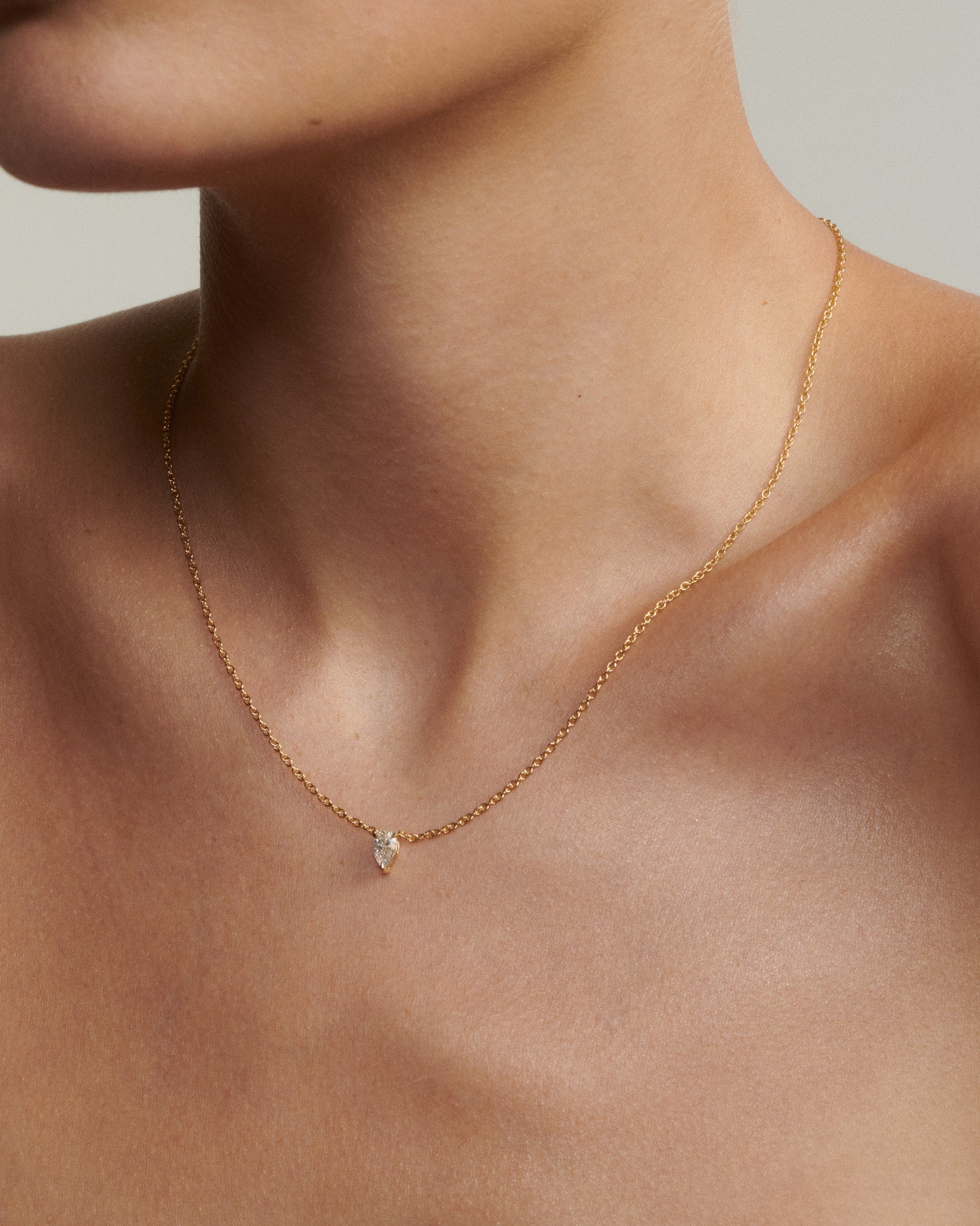 Gold necklace with a small pendant on a person's neck against a neutral background [text overlay: Andrea wears the 0.5ct Pear Diamond Necklace]