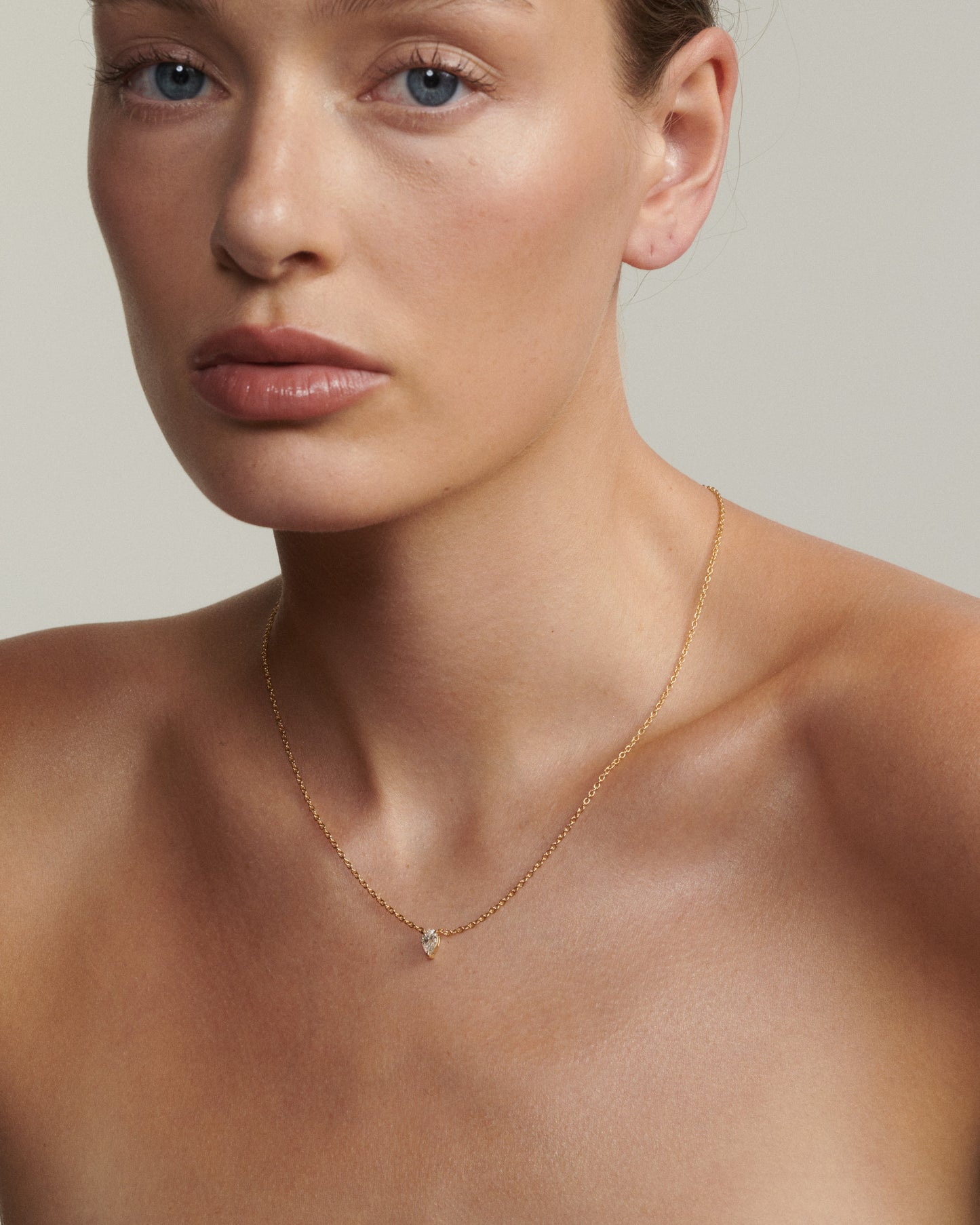 Woman wearing a delicate gold necklace with a small pendant against a neutral background [text overlay: Andrea wears the 0.5ct Pear Diamond Necklace]
