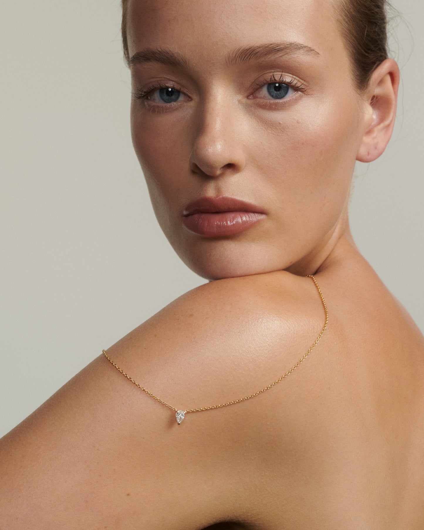 Woman wearing a delicate gold necklace with a small diamond pendant on a neutral background [text overlay: Andrea wears the 0.5ct Pear Diamond Necklace]