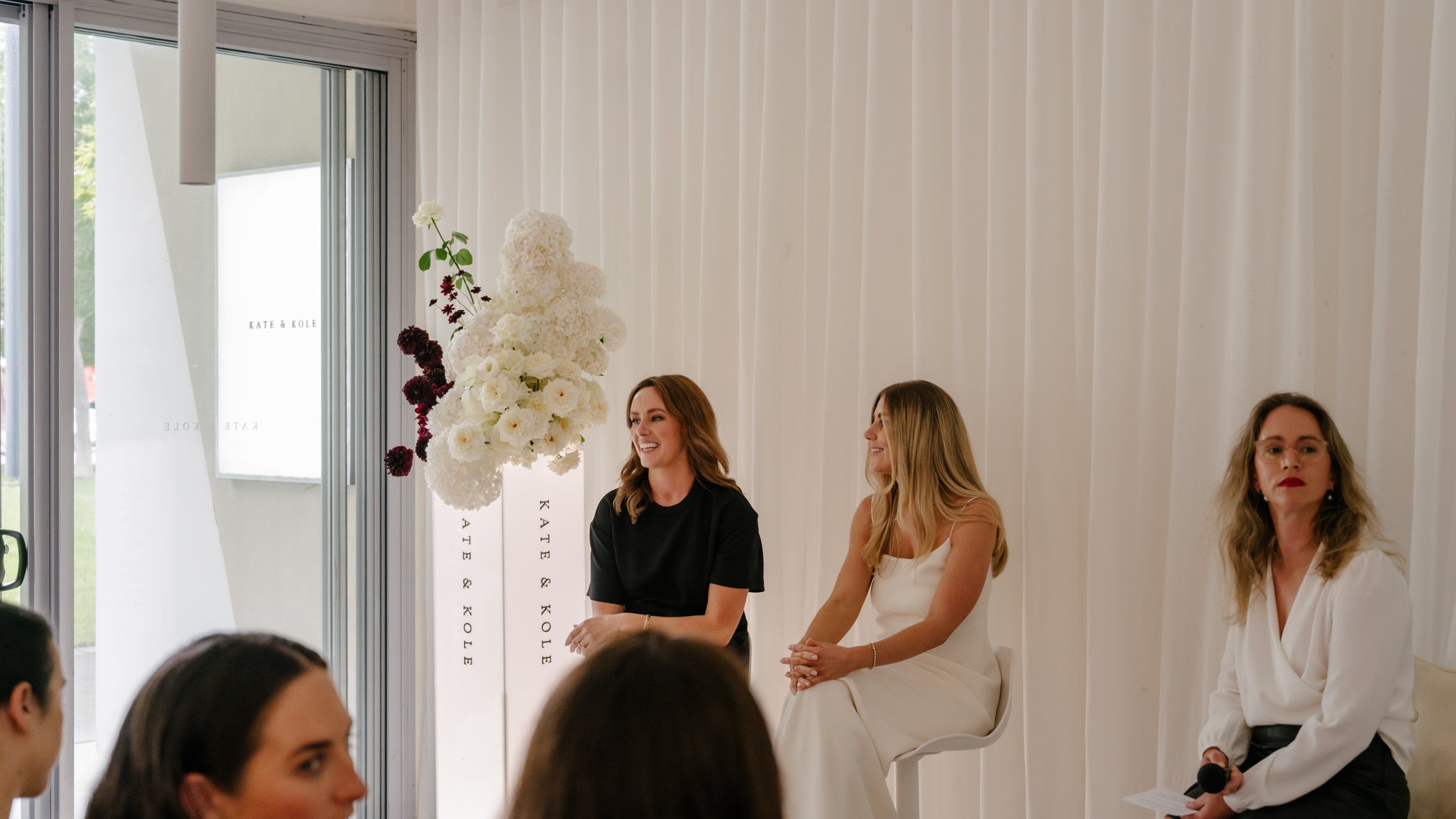 Women in elegant attire at a luxury jewellery event, seated against a white backdrop with floral arrangements and signage.