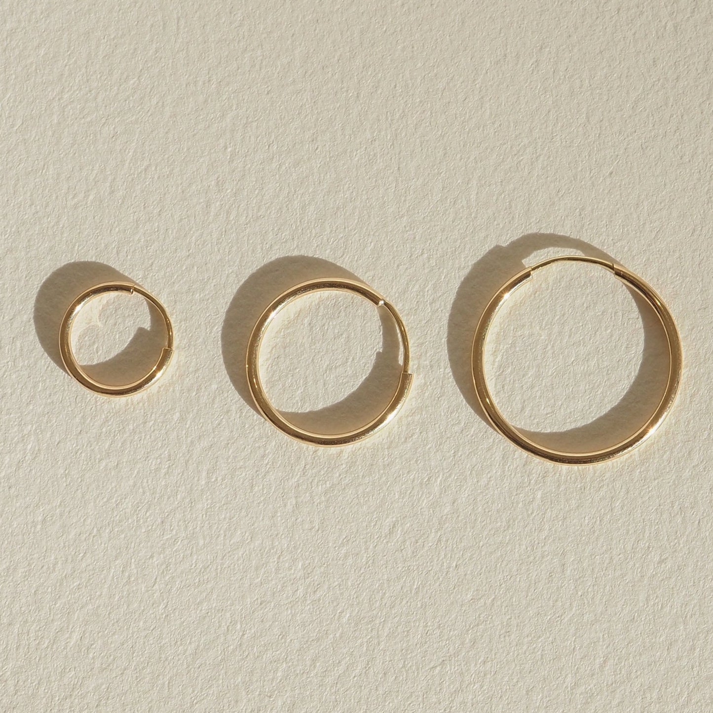 Four gold bangles of varying sizes displayed on a light textured surface, casting soft shadows, showcasing minimalist luxury jewellery design.